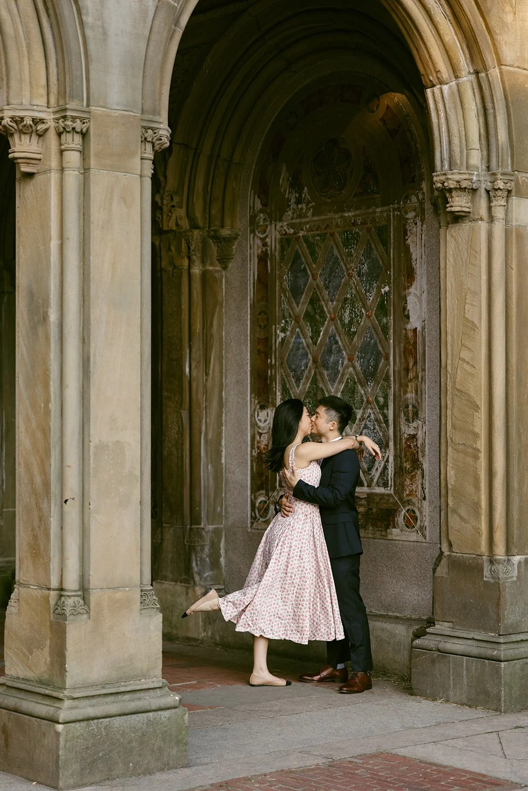 A couple embraces and kisses under a stone archway with ornate detailing and a patterned wall in the background.