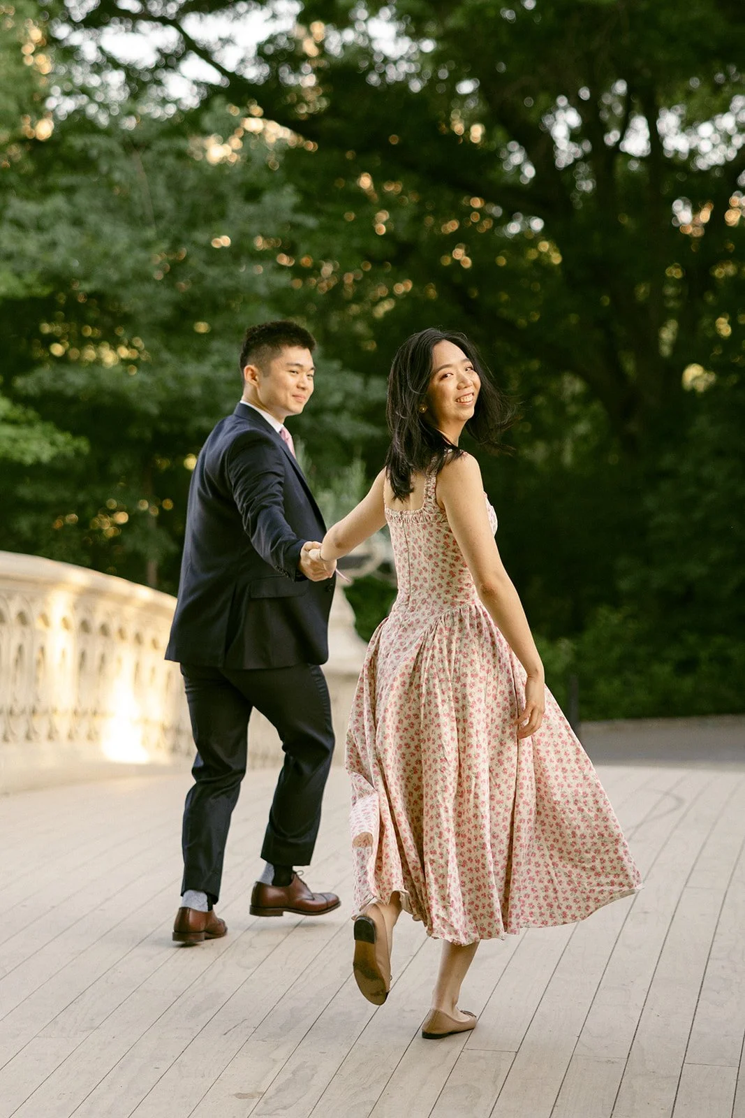 A woman in a floral dress leads a man in a suit by the hand across a wooden bridge, with trees in the background for central park engagement photos