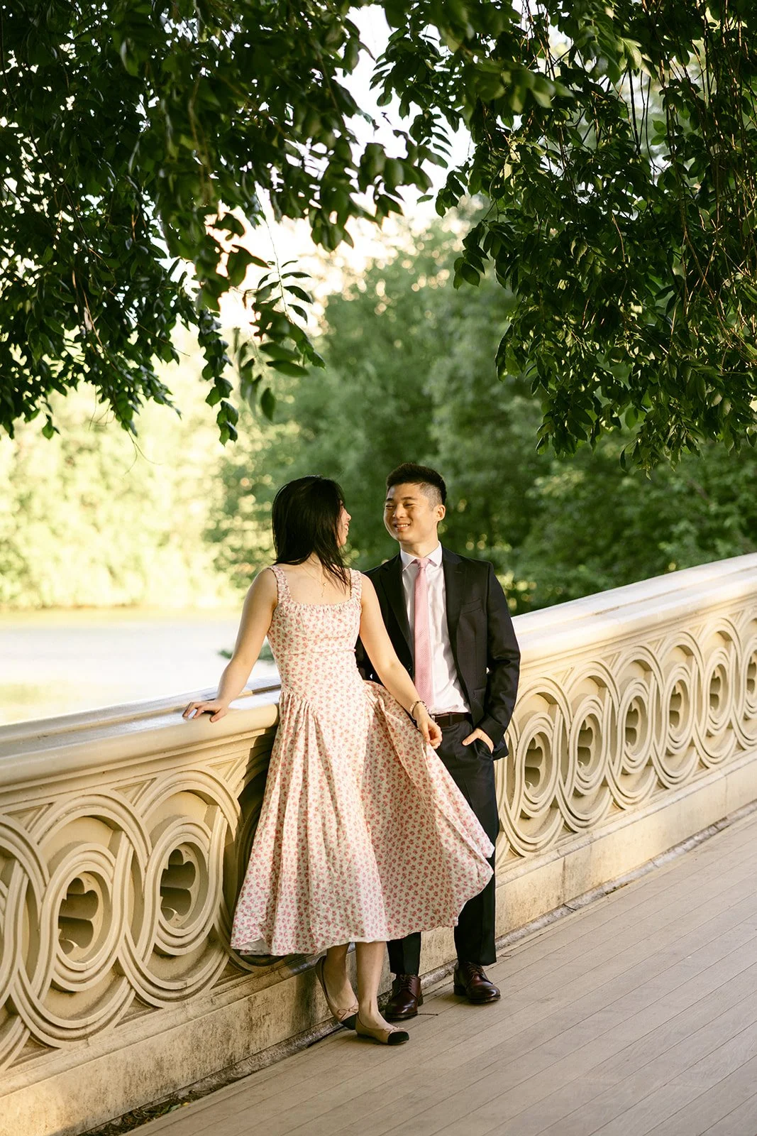 A woman in a floral dress leads a man in a suit by the hand across a wooden bridge, with trees in the background for central park engagement photos