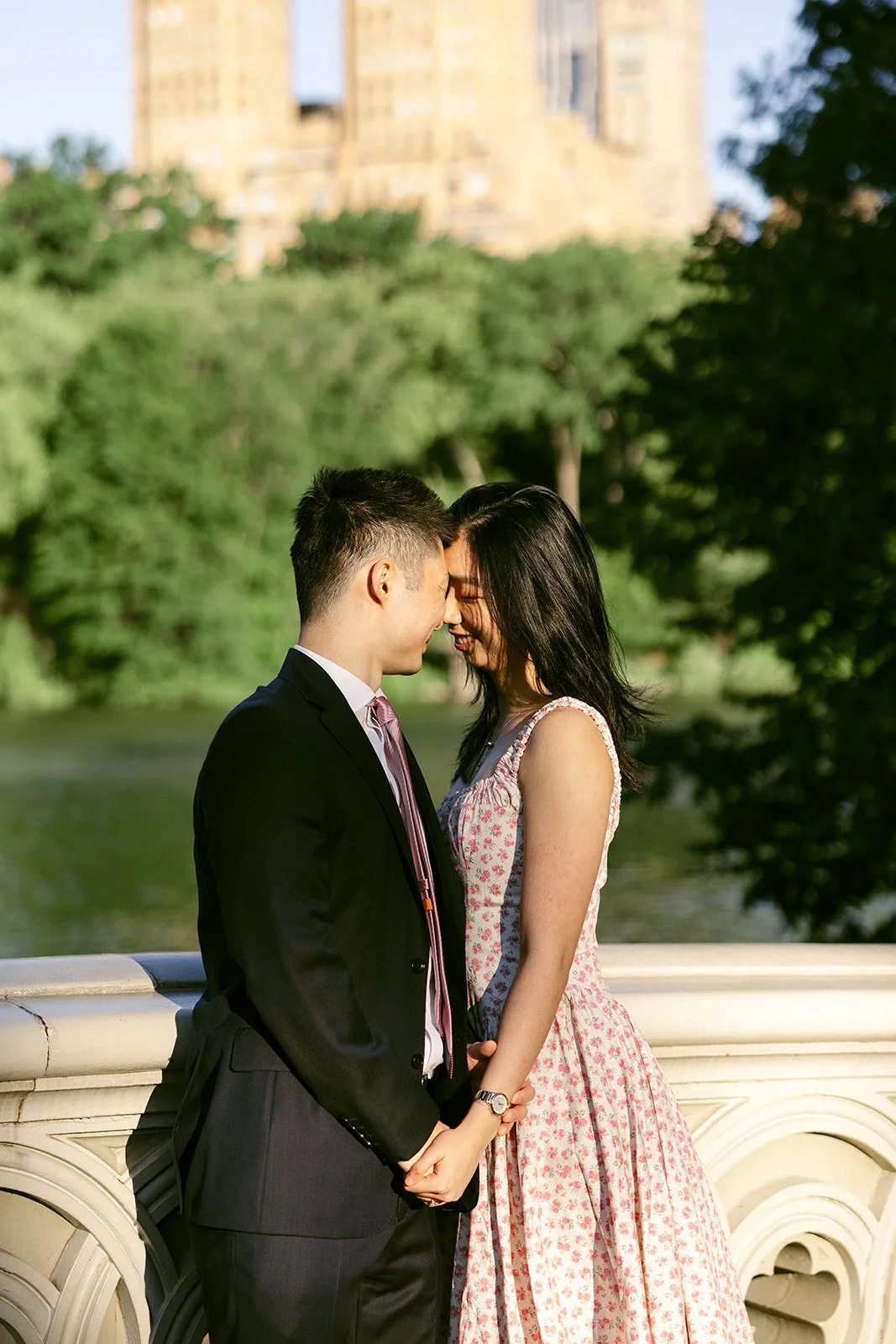 A couple stands close together on a bridge, out of focus, with tall buildings, trees, and a body of water in the background on a clear day.