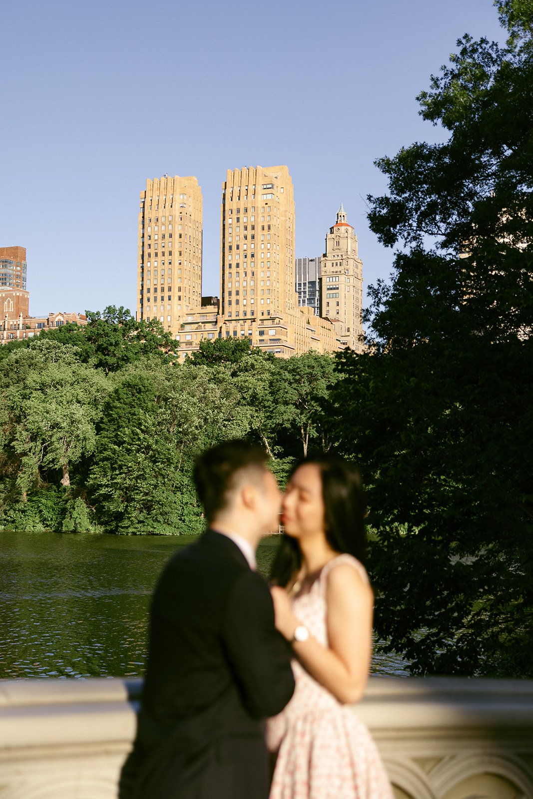 A couple stands close together on a bridge, out of focus, with tall buildings, trees, and a body of water in the background on a clear day.