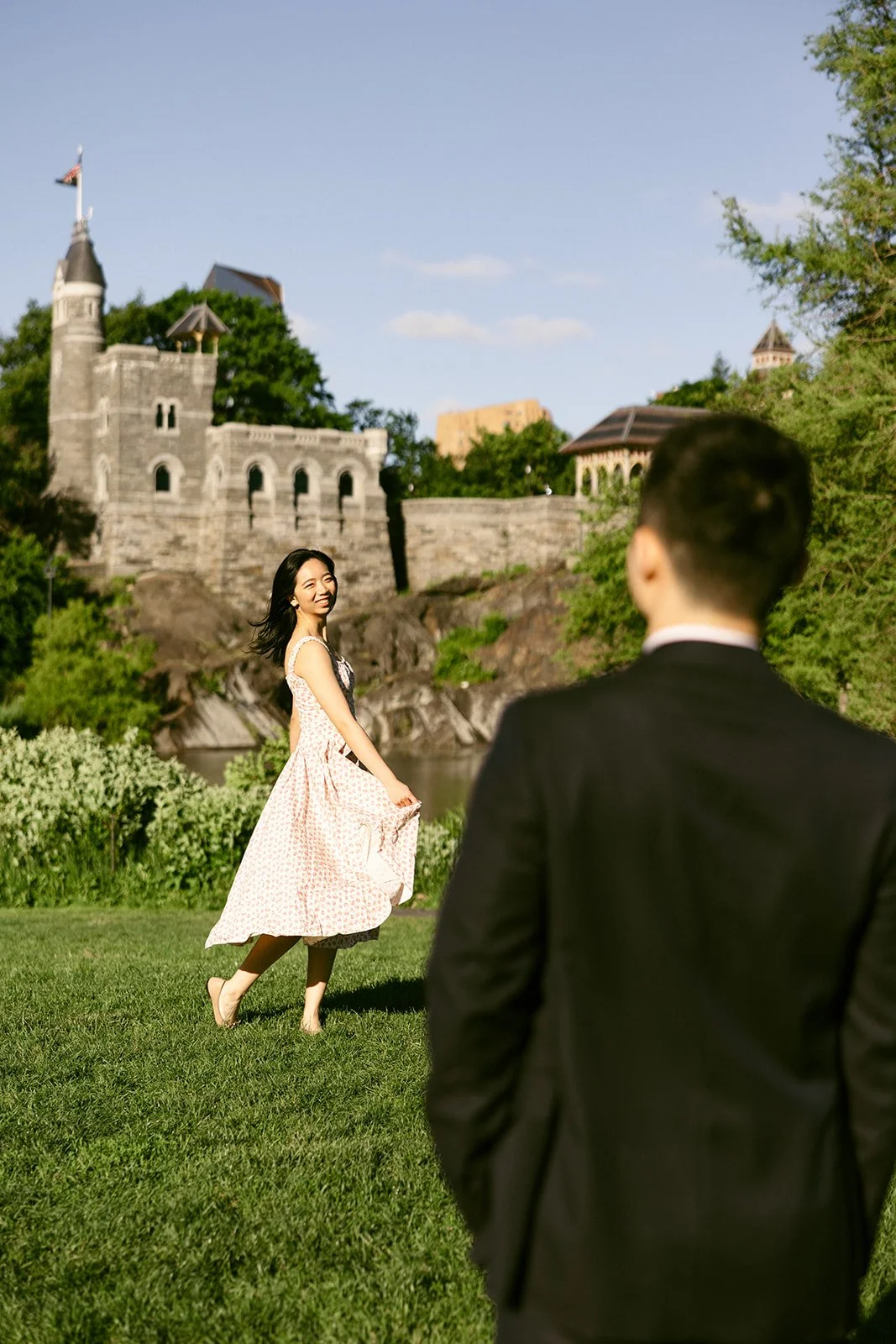A woman in a light dress spins on grass while a man in a suit watches her; a stone building and trees are in the background under a clear sky foe central park engagement photos 