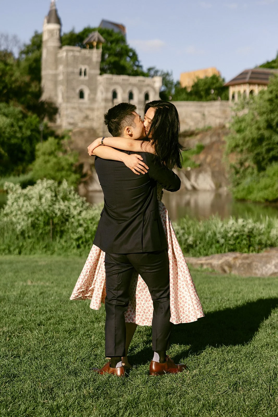 A woman in a light dress spins on grass while a man in a suit watches her; a stone building and trees are in the background under a clear sky foe central park engagement photos 