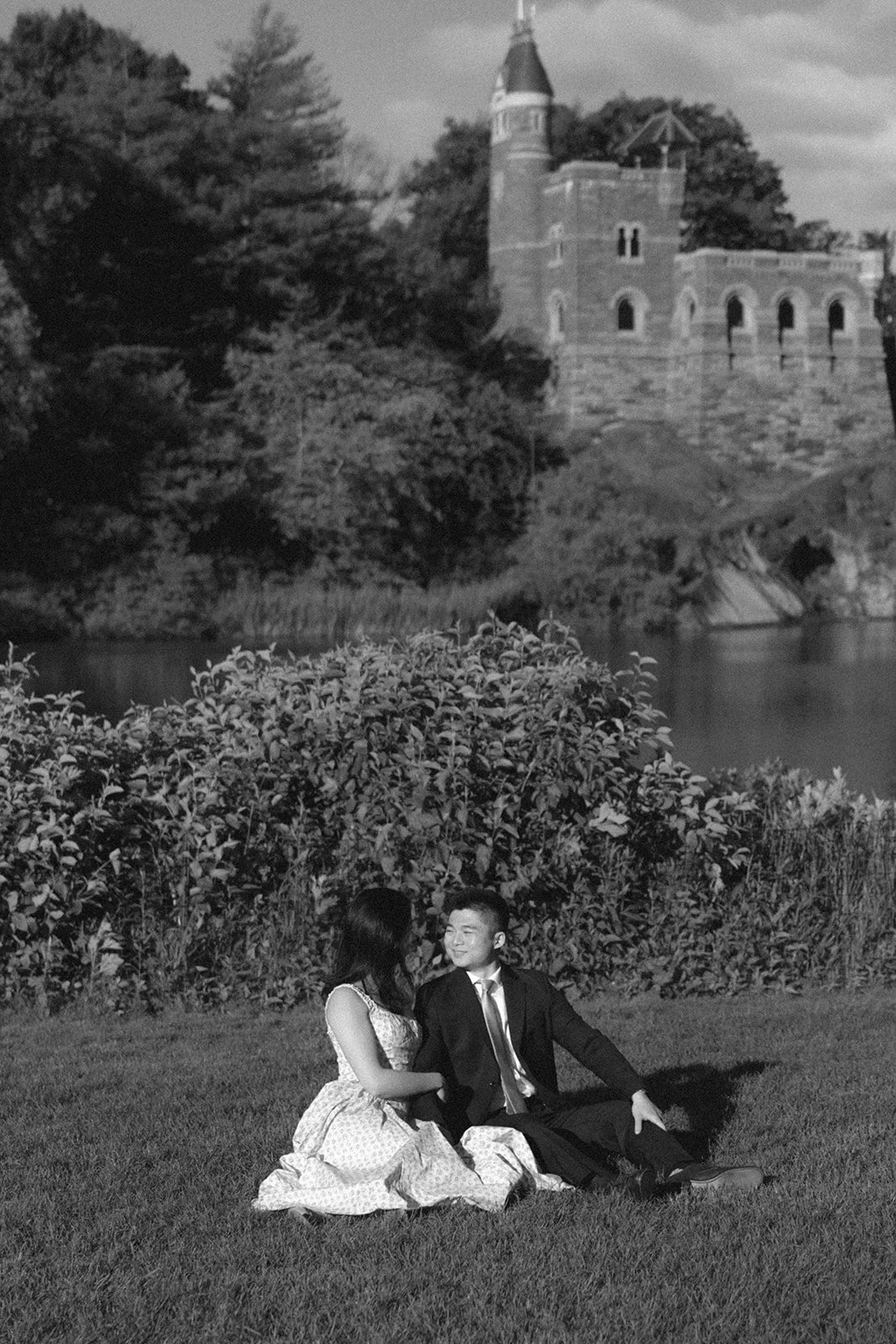 A woman in a light dress spins on grass while a man in a suit watches her; a stone building and trees are in the background under a clear sky for central park engagement photos 