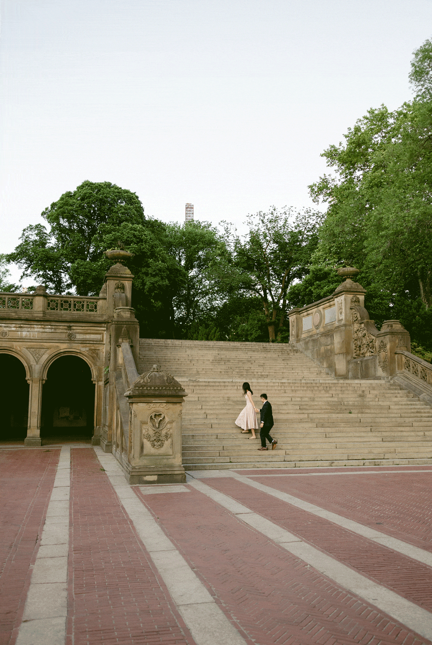 A couple in formal attire walks up stone steps in an outdoor plaza surrounded by trees and historic architecture.