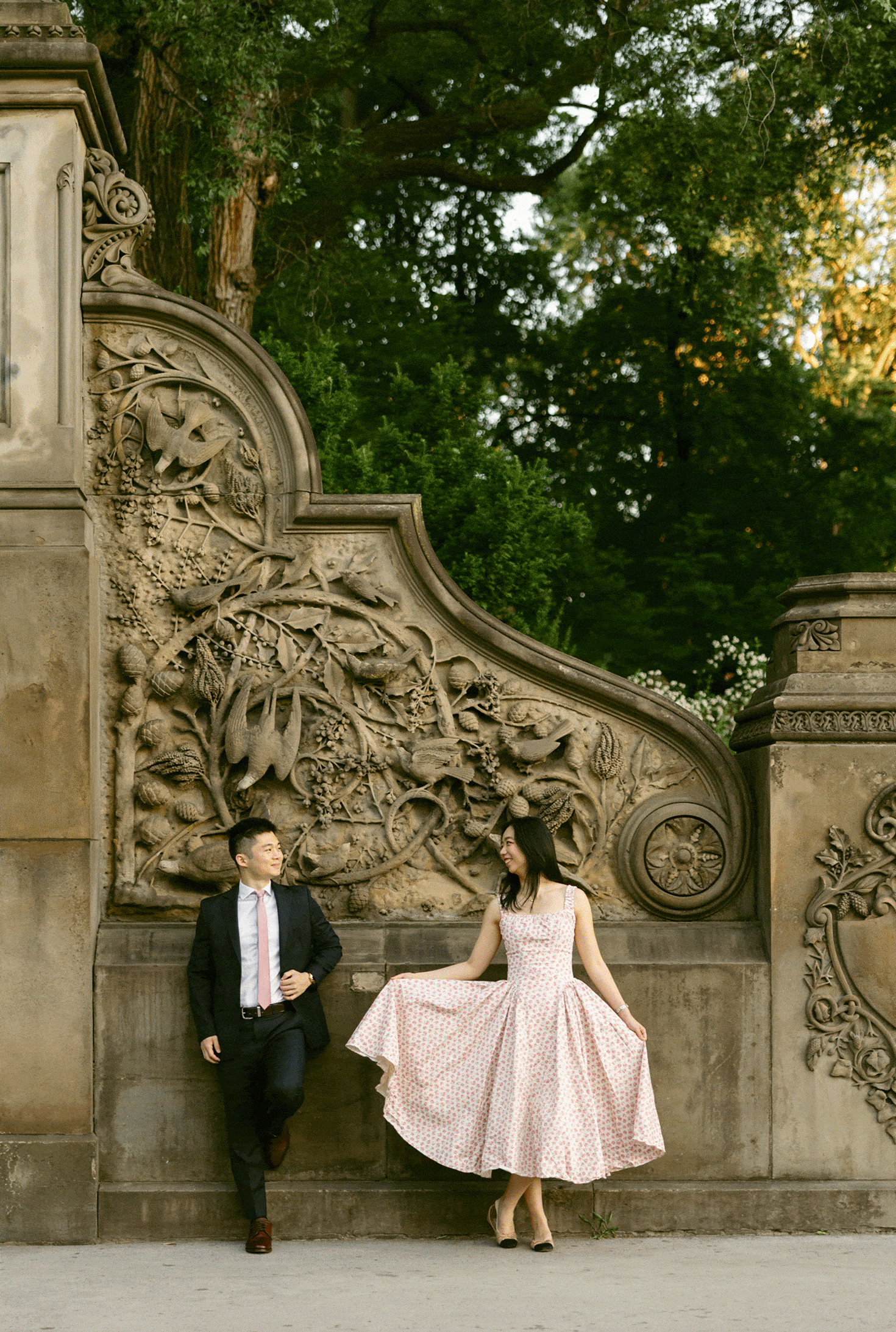 A man in a suit and a woman in a patterned dress stand by an ornate stone wall in an outdoor setting with greenery.