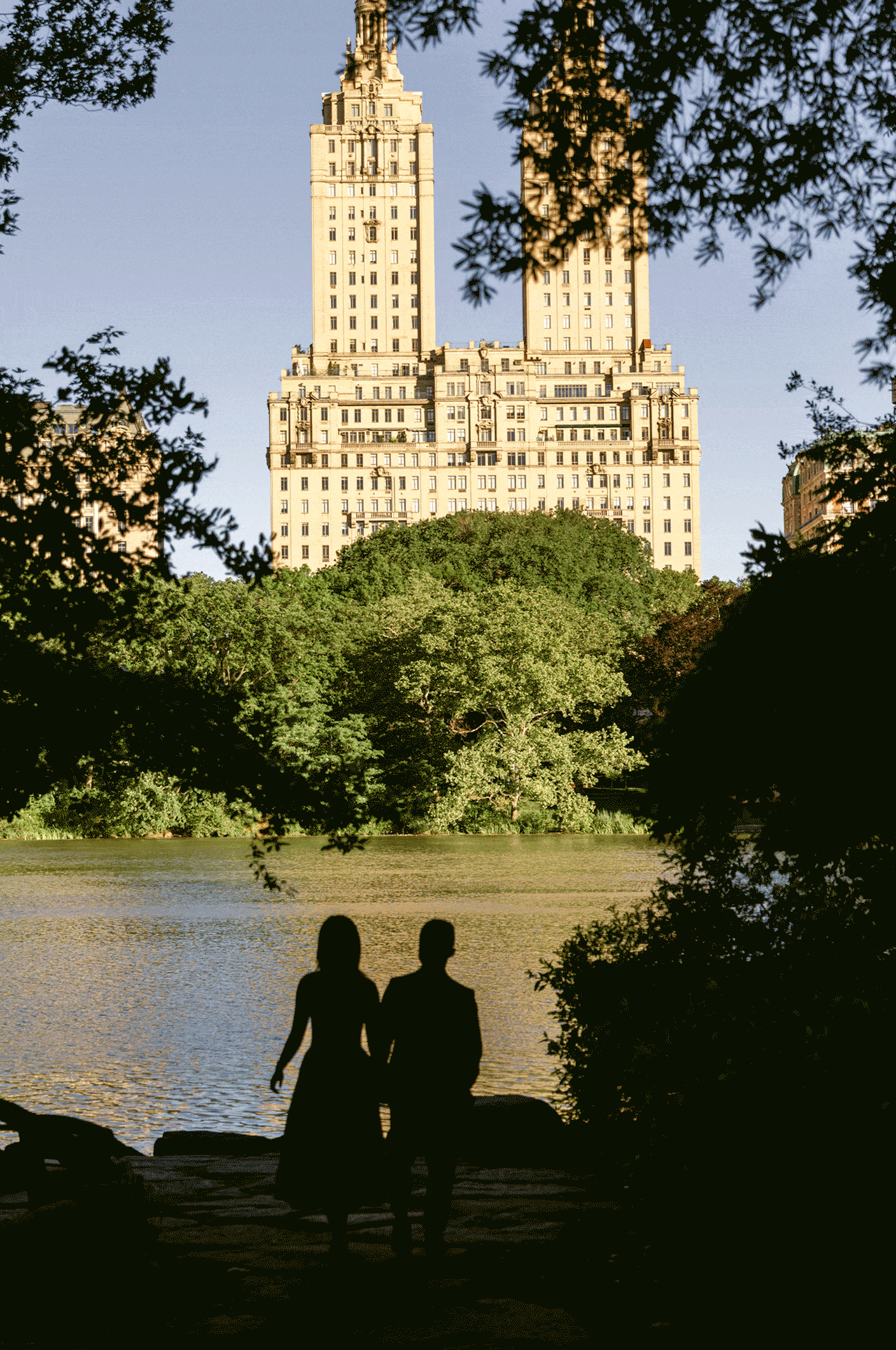 Two people stand by a lake with tall buildings and trees in the background, framed by tree branches.