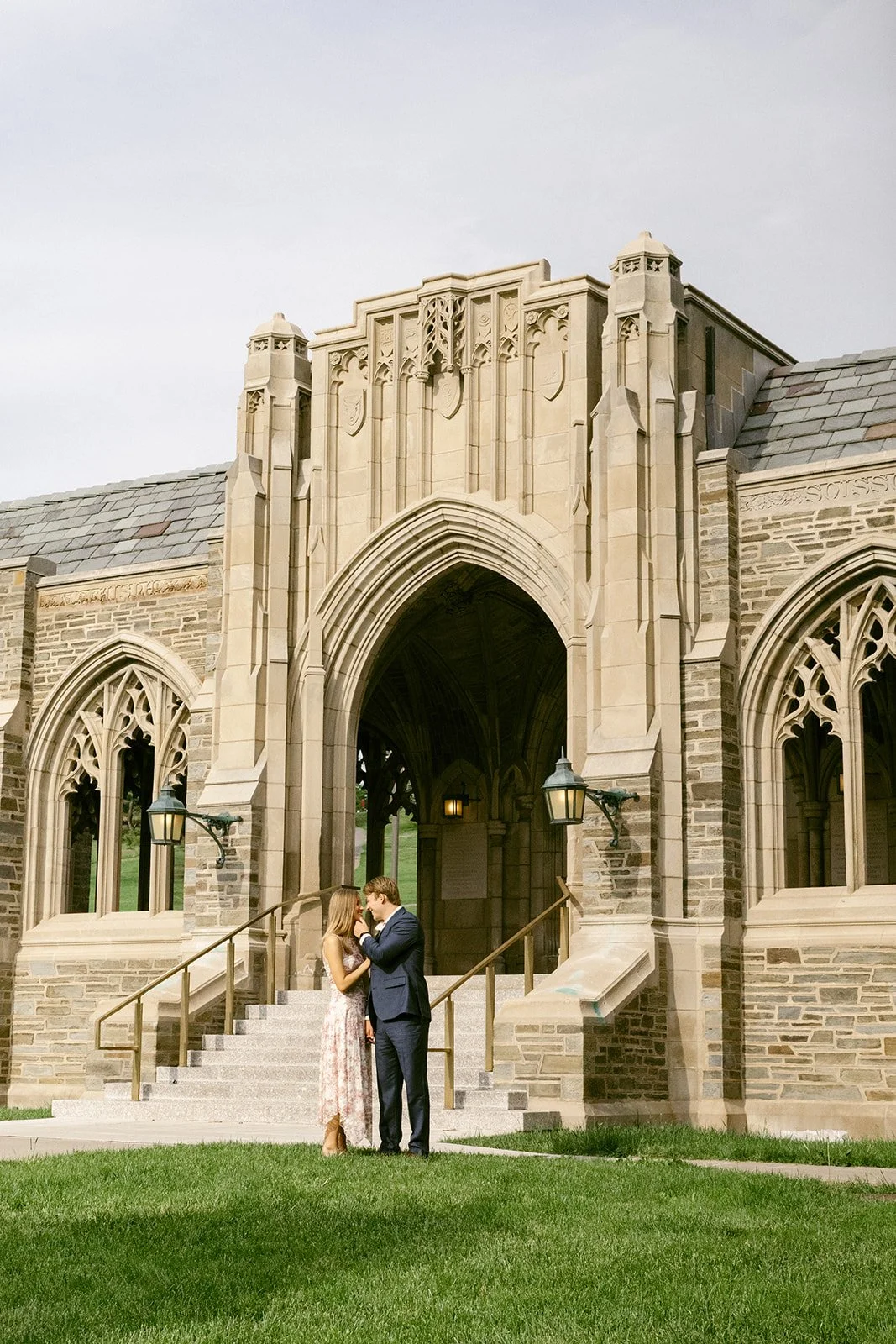 A couple stands embraced on the steps of a large stone building for outdoor engagement photos at cornell university