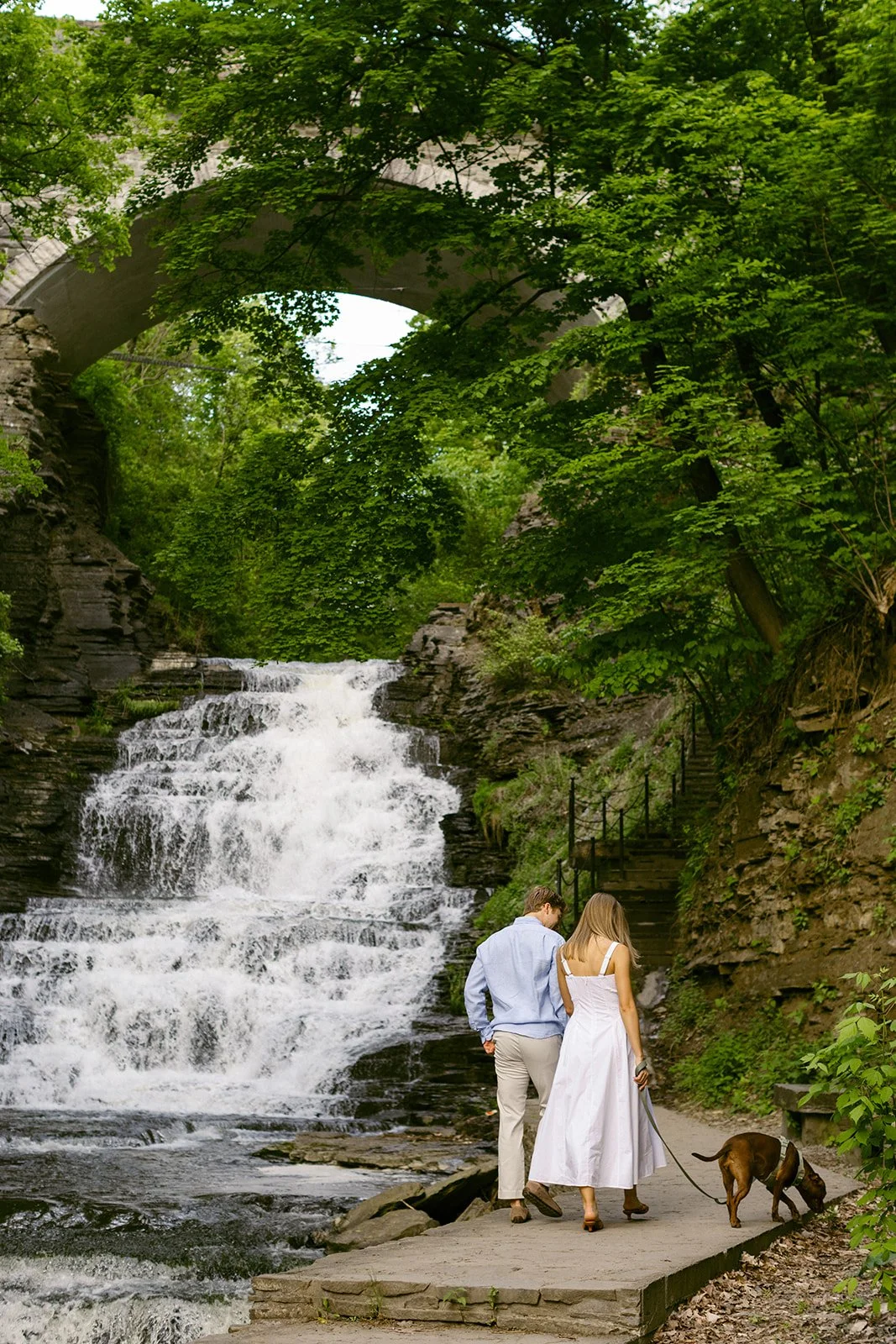 A couple walks a dog along a path next to a waterfall, with a stone bridge and lush green trees in the background for outdoor engagement photos