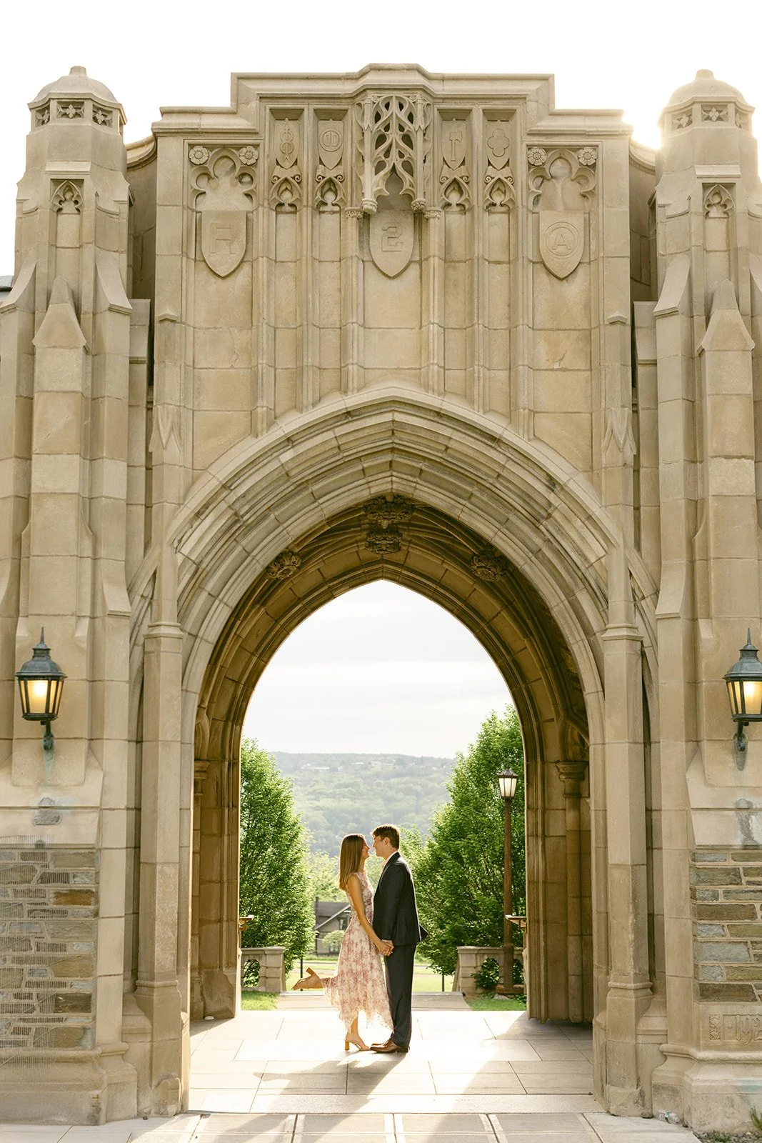 A couple stands embraces in front of a large stone building for outdoor engagement photos at cornell university