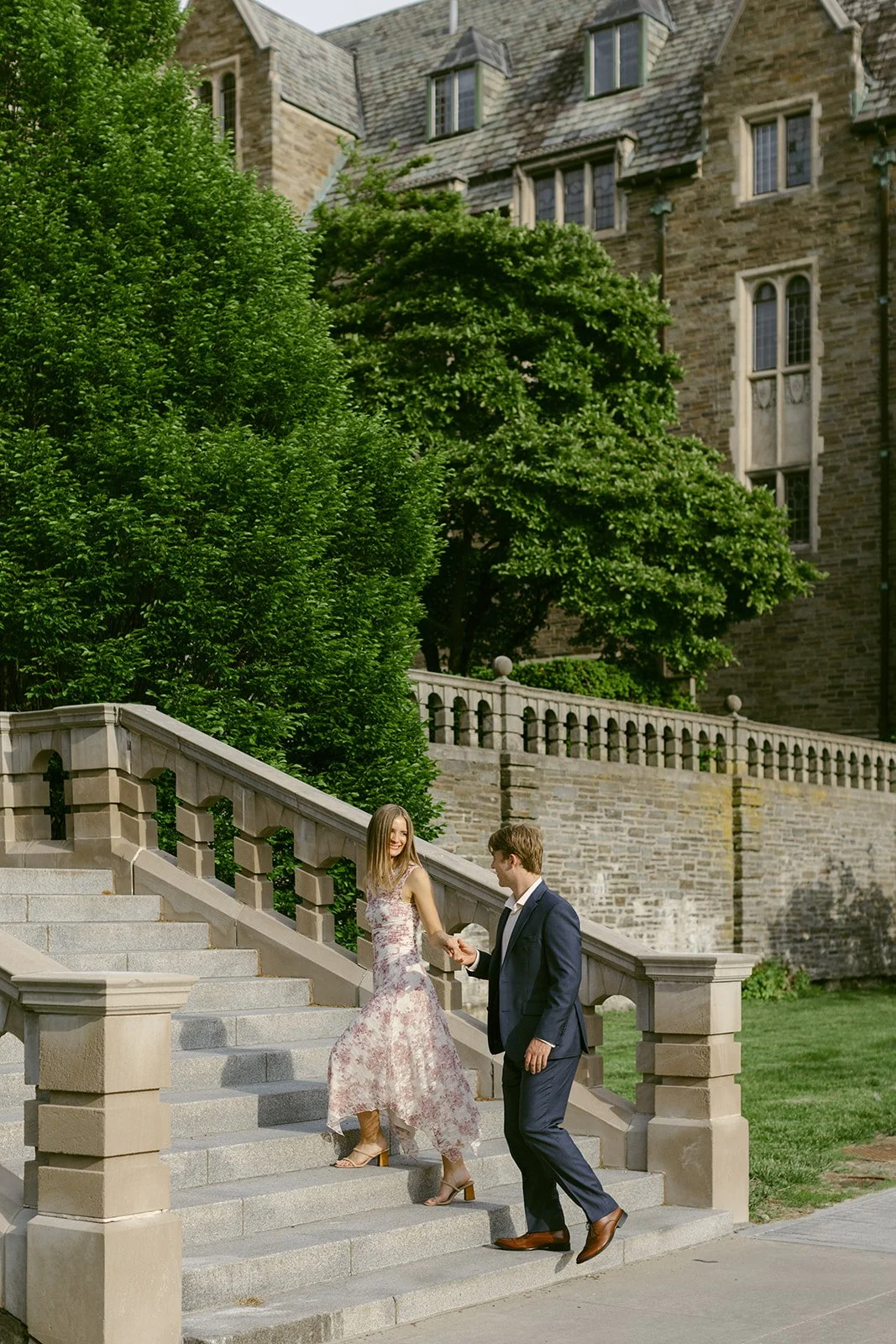 A woman in a floral dress holds hands with a man in a dark suit as they walk down stone steps outside cornell university for outdoor engagement photos 