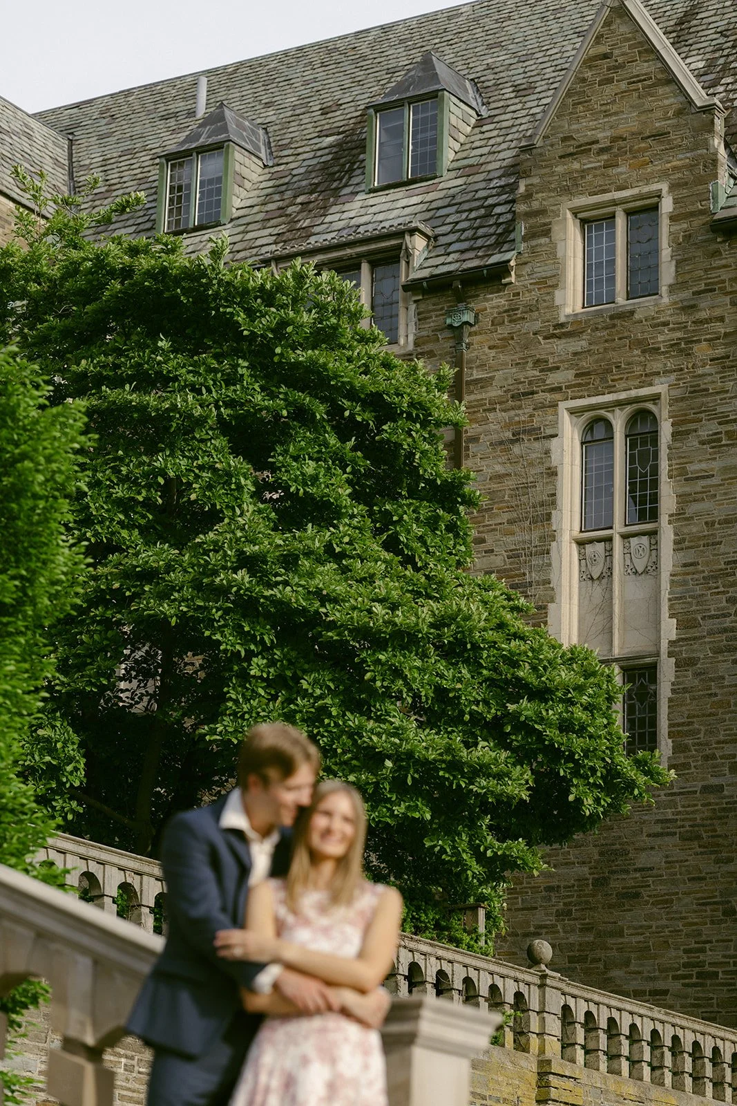A woman in a floral dress holds hands with a man in a dark suit as they walk down stone steps outside cornell university for outdoor engagement photos 