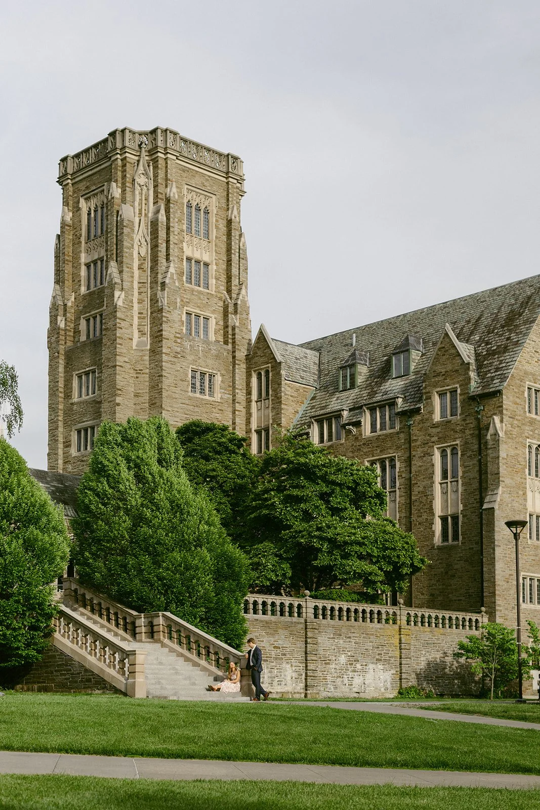 A woman in a floral dress holds hands with a man in a dark suit as they walk down stone steps outside cornell university for outdoor engagement photos 
