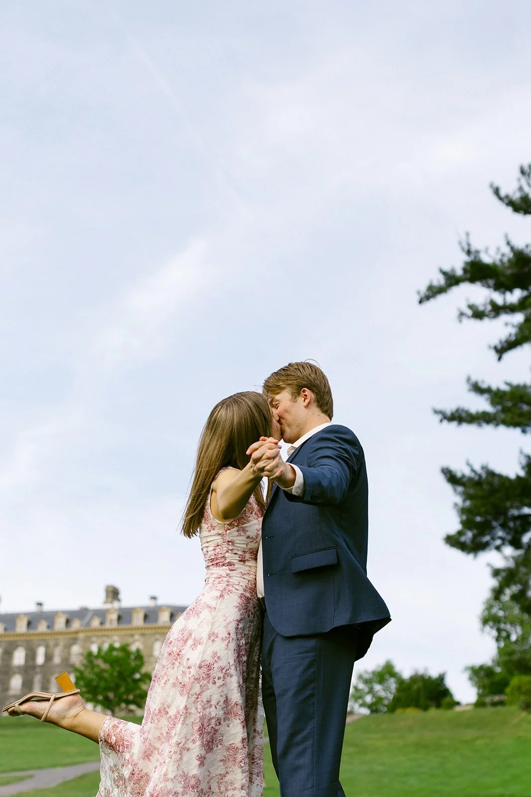 A man in a suit and a woman in a dress walk hand in hand across a grassy lawn toward a large stone building on a partly cloudy day.