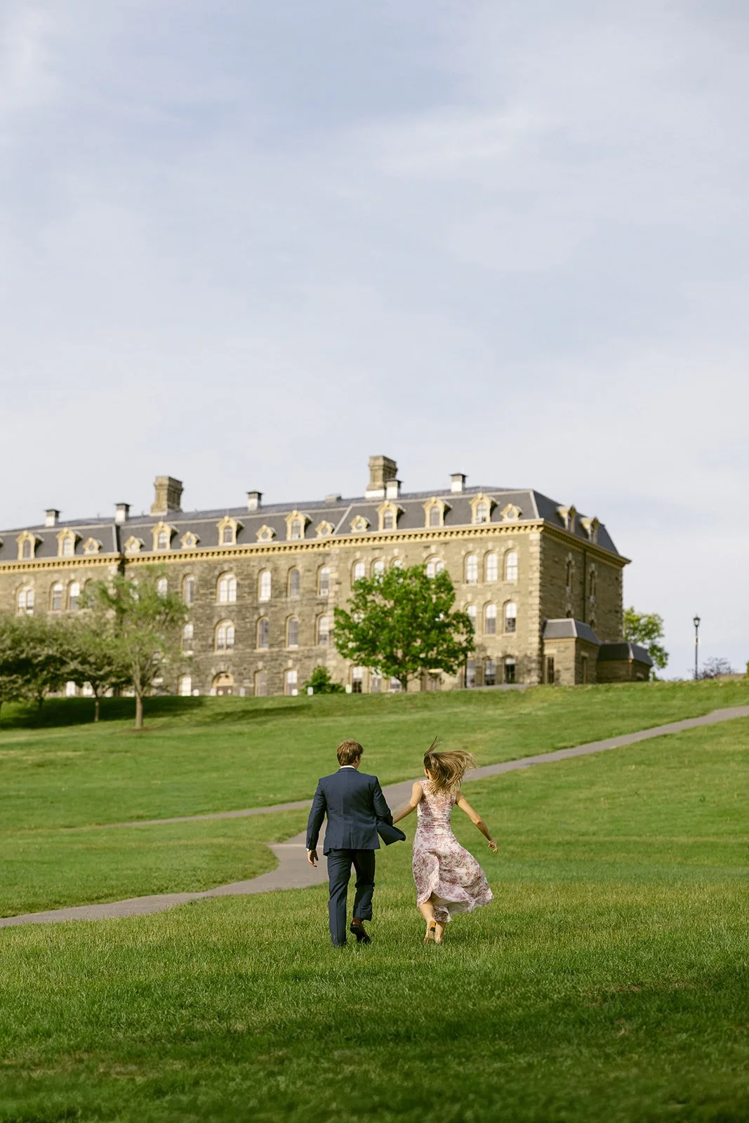 A man in a suit and a woman in a dress walk hand in hand across a grassy lawn toward a large stone building on a partly cloudy day.