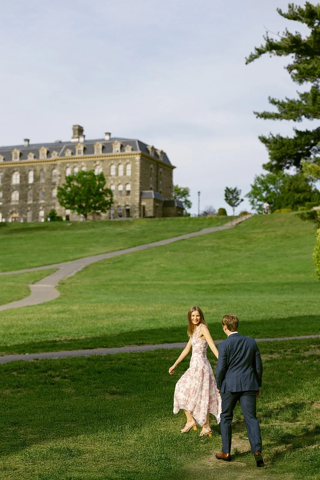 A man in a suit and a woman in a dress walk hand in hand across a grassy lawn toward a large stone building on a partly cloudy day.