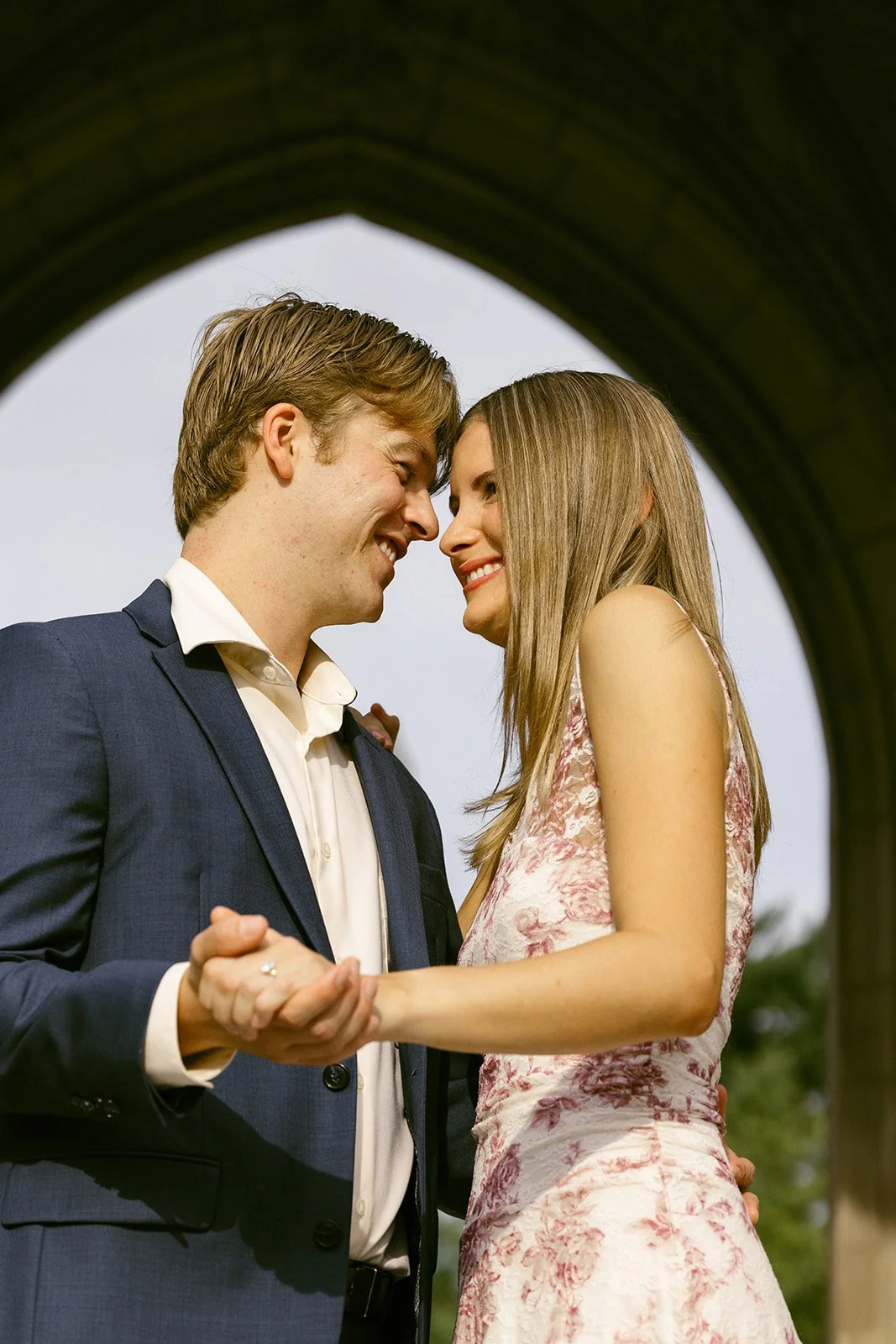 A man in a suit and a woman in a floral dress smile at each other while holding hands under a large arch outdoors.