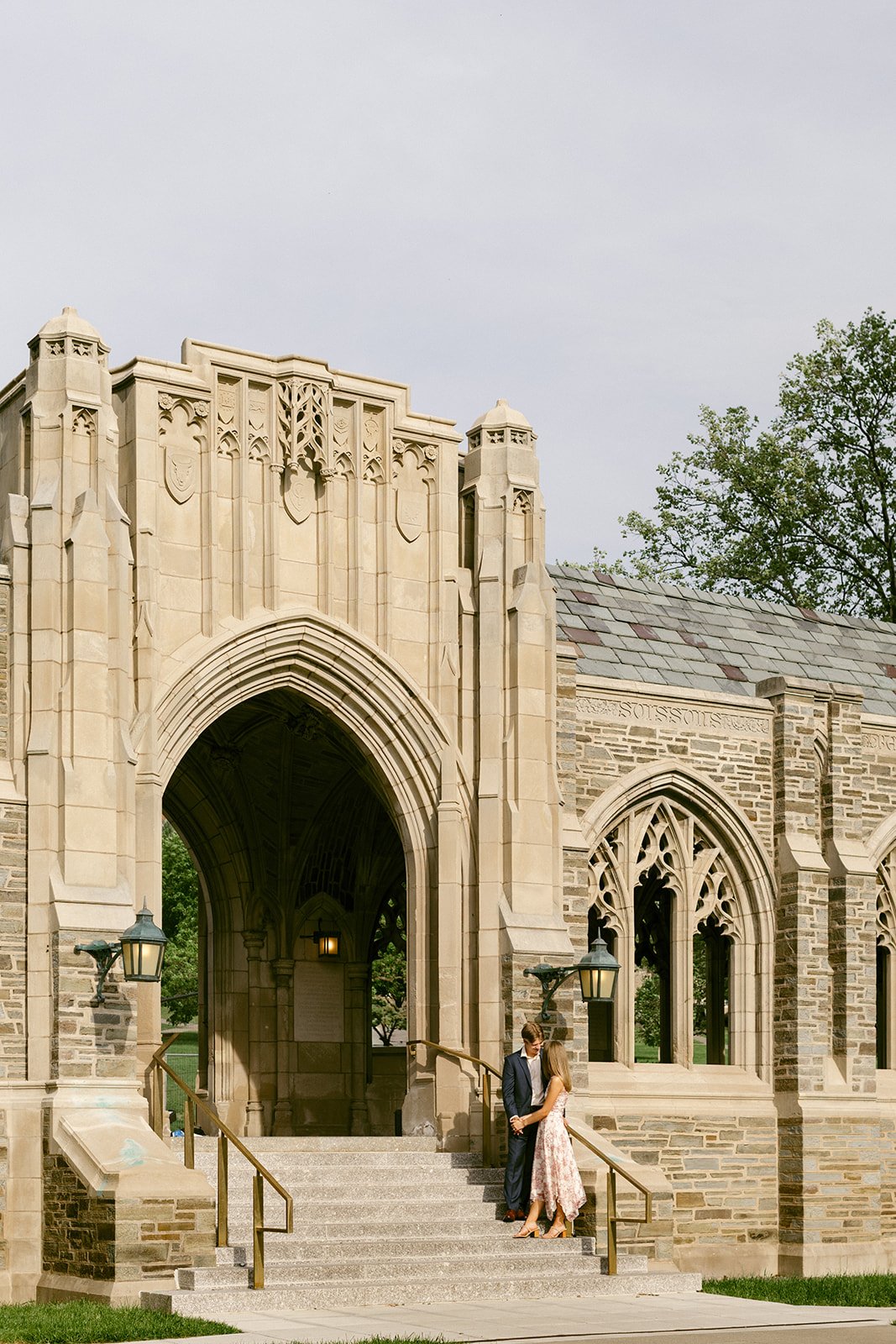 A couple stands embraced on the steps of a large stone building for outdoor engagement photos at cornell university