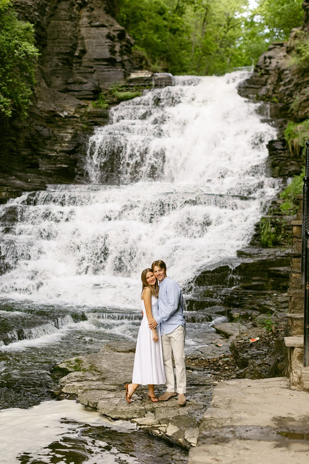 A couple stands together at the base of a multi-tiered waterfall surrounded by greenery and rocky cliffs, with water cascading down behind them for outdoor engagement photos at cascadilla gorge