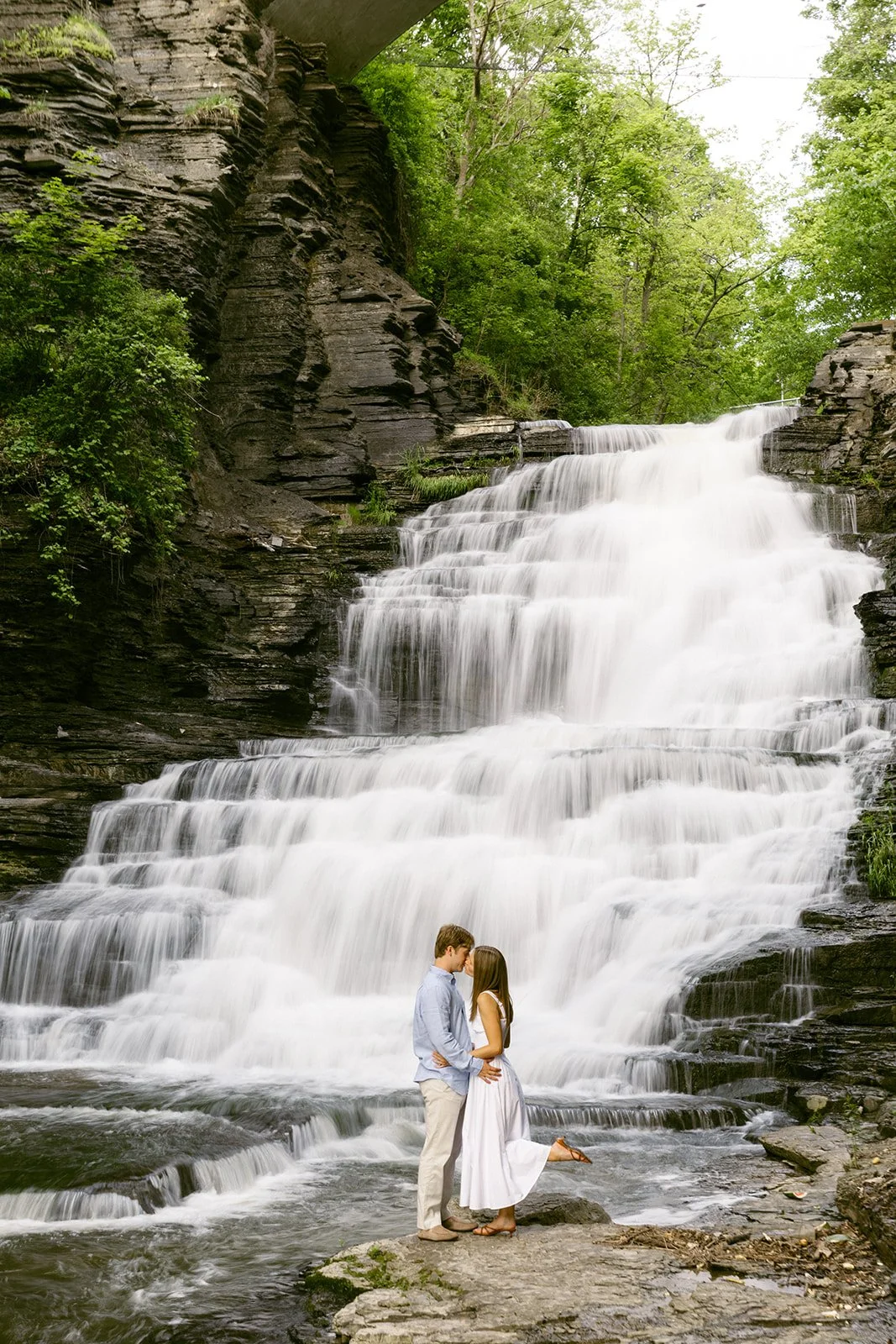 A couple stands together at the base of a multi-tiered waterfall surrounded by greenery and rocky cliffs, with water cascading down behind them for outdoor engagement photos at cascadilla gorge