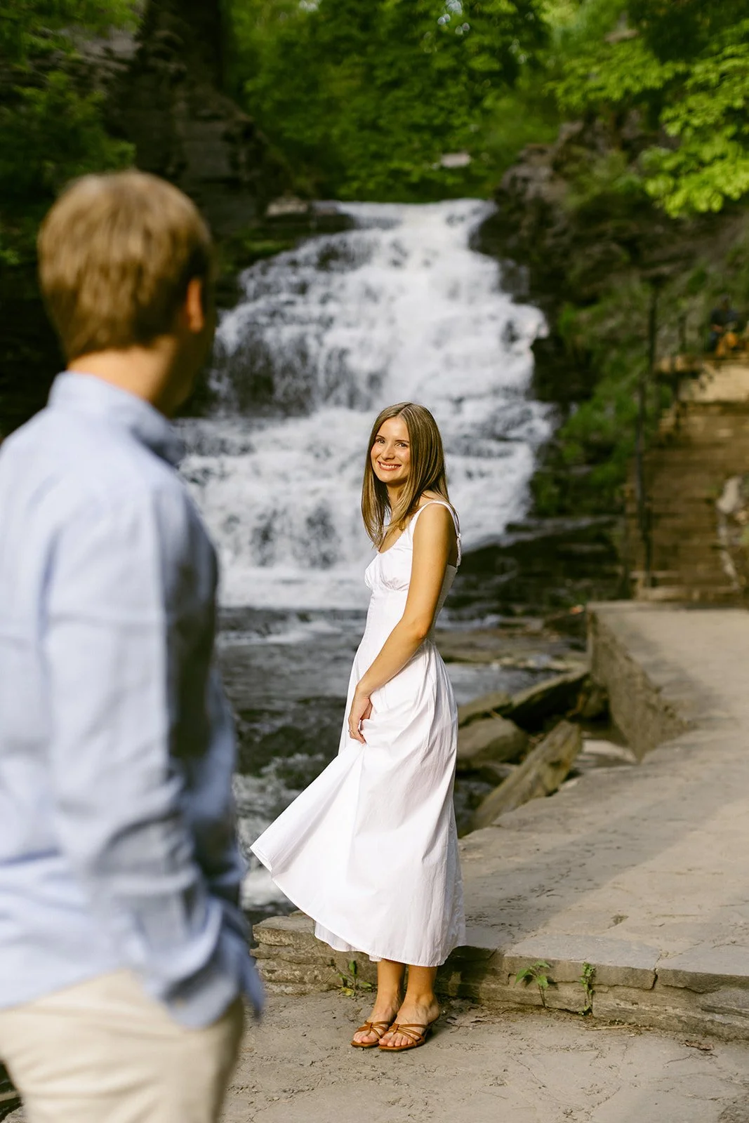A couple stands together at the base of a multi-tiered waterfall surrounded by greenery and rocky cliffs, with water cascading down behind them for outdoor engagement photos at cascadilla gorge