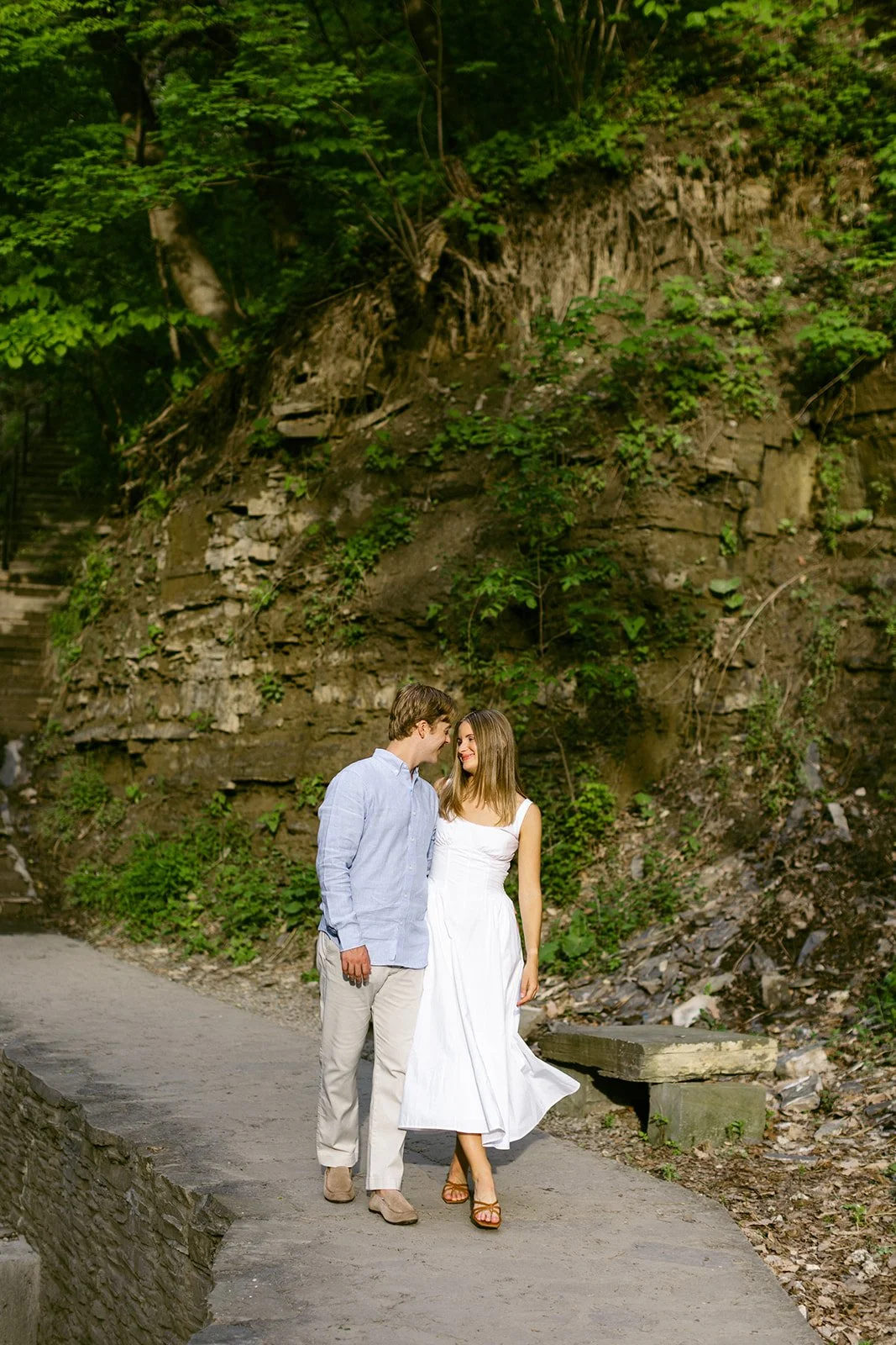 A couple stands together at the base of a multi-tiered waterfall surrounded by greenery and rocky cliffs, with water cascading down behind them for outdoor engagement photos at cascadilla gorge