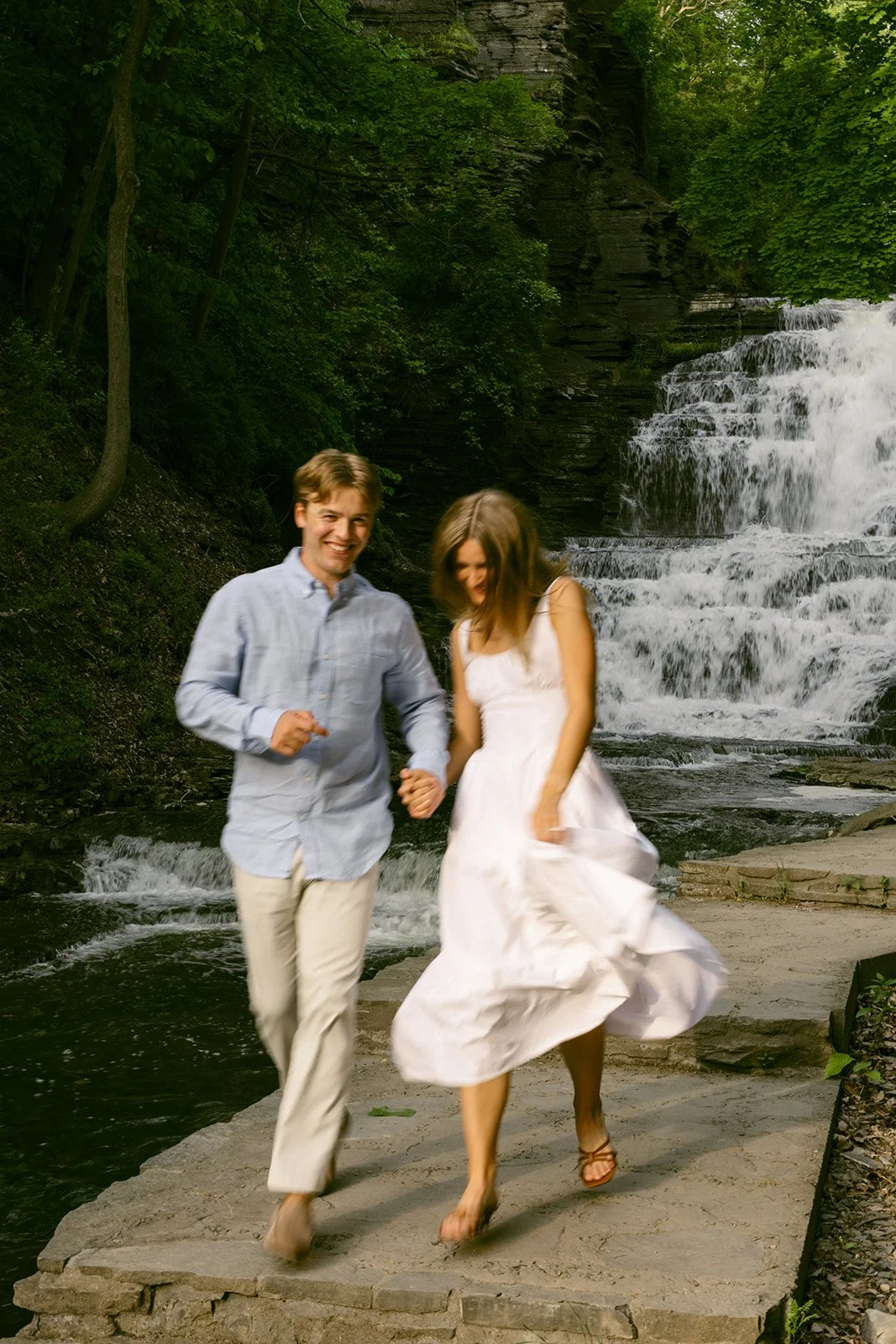 A couple stands together at the base of a multi-tiered waterfall surrounded by greenery and rocky cliffs, with water cascading down behind them for outdoor engagement photos at cascadilla gorge