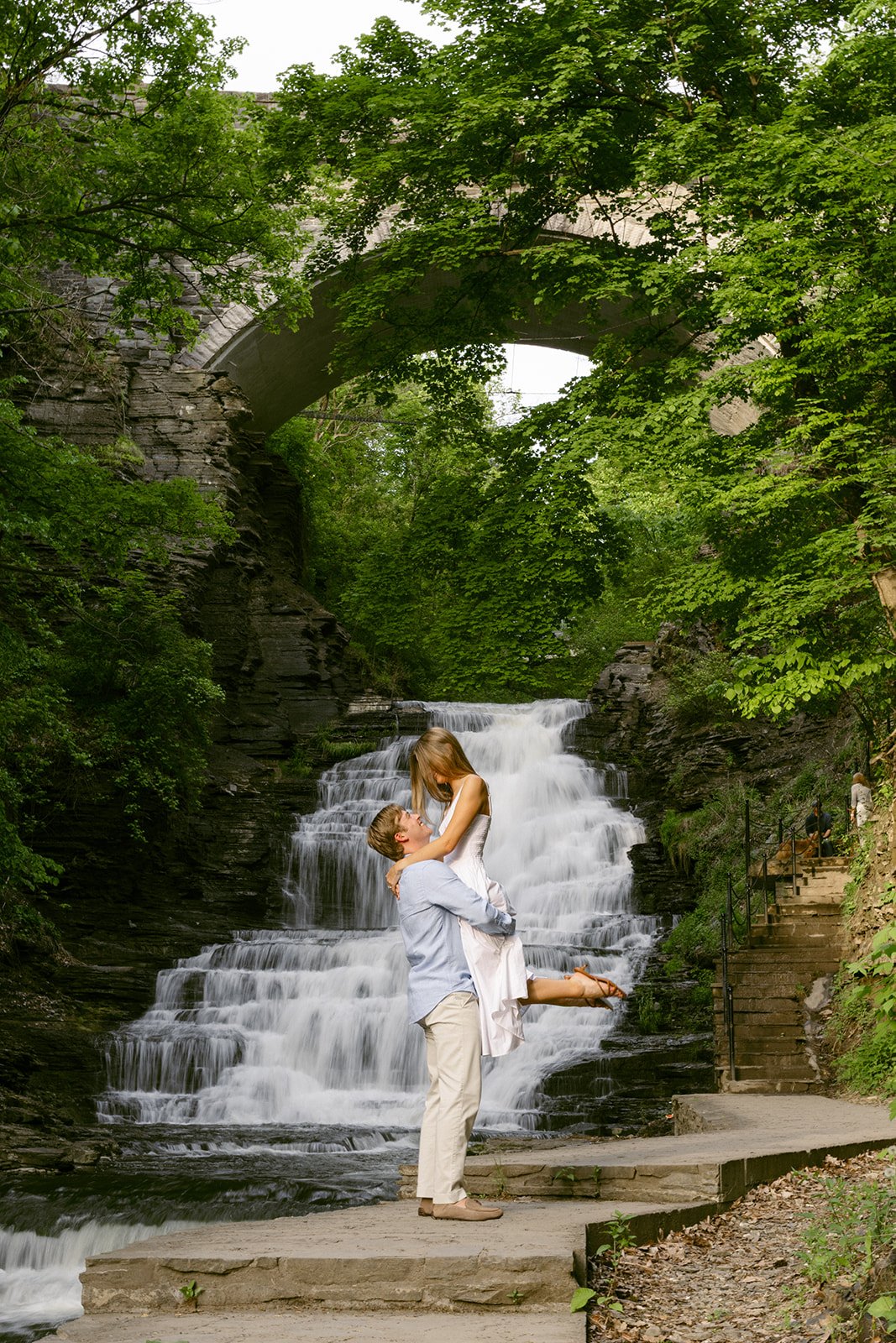 A couple stands together at the base of a multi-tiered waterfall surrounded by greenery and rocky cliffs, with water cascading down behind them for outdoor engagement photos at cascadilla gorge