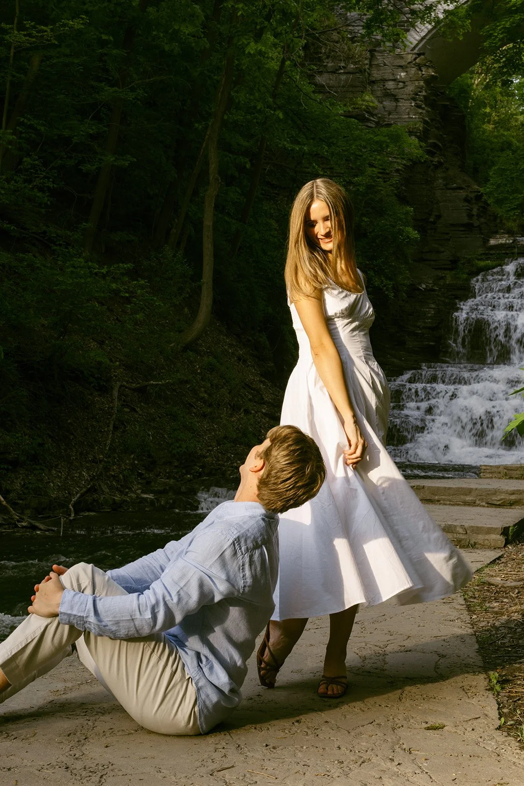 A couple stands together at the base of a multi-tiered waterfall surrounded by greenery and rocky cliffs, with water cascading down behind them for outdoor engagement photos at cascadilla gorge