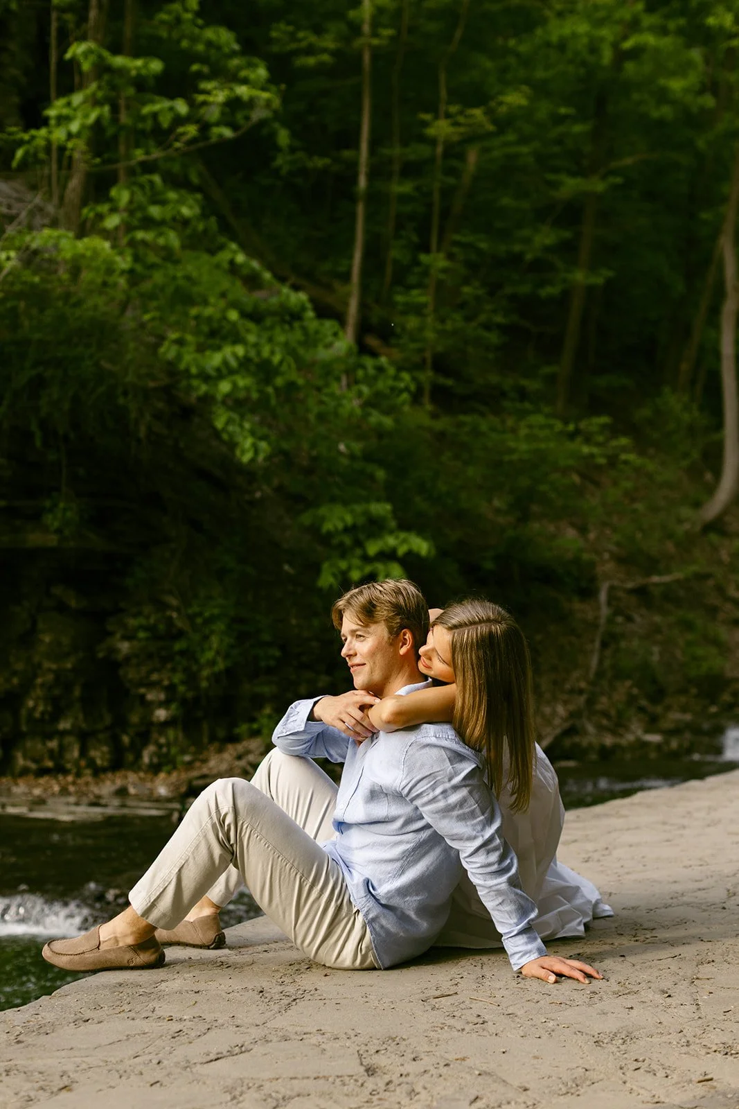 A couple stands together at the base of a multi-tiered waterfall surrounded by greenery and rocky cliffs, with water cascading down behind them for outdoor engagement photos at cascadilla gorge
