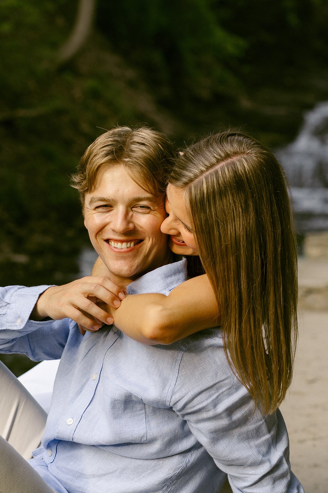 A couple stands together at the base of a multi-tiered waterfall surrounded by greenery and rocky cliffs, with water cascading down behind them for outdoor engagement photos at cascadilla gorge