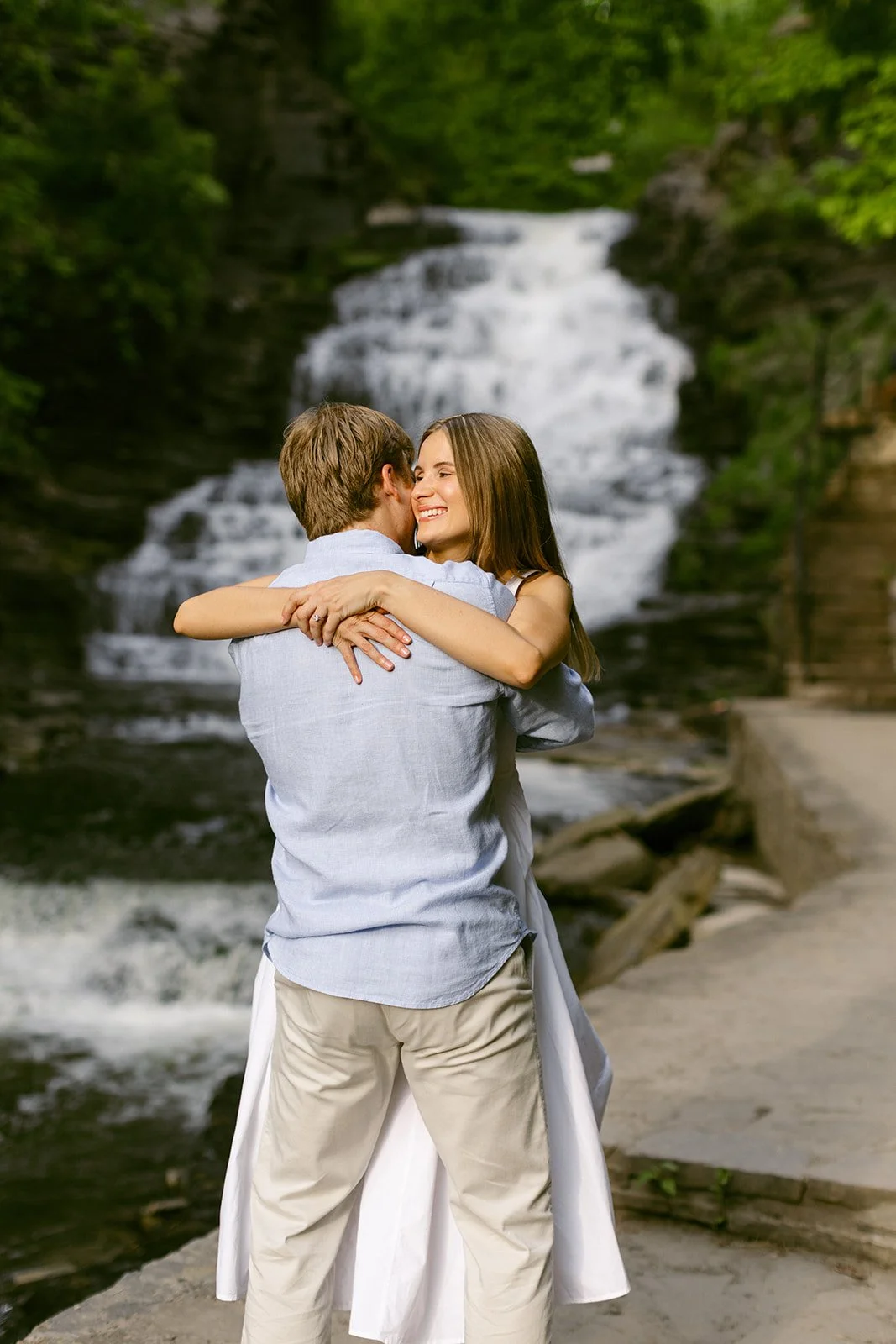 A couple stands together at the base of a multi-tiered waterfall surrounded by greenery and rocky cliffs, with water cascading down behind them for outdoor engagement photos at cascadilla gorge