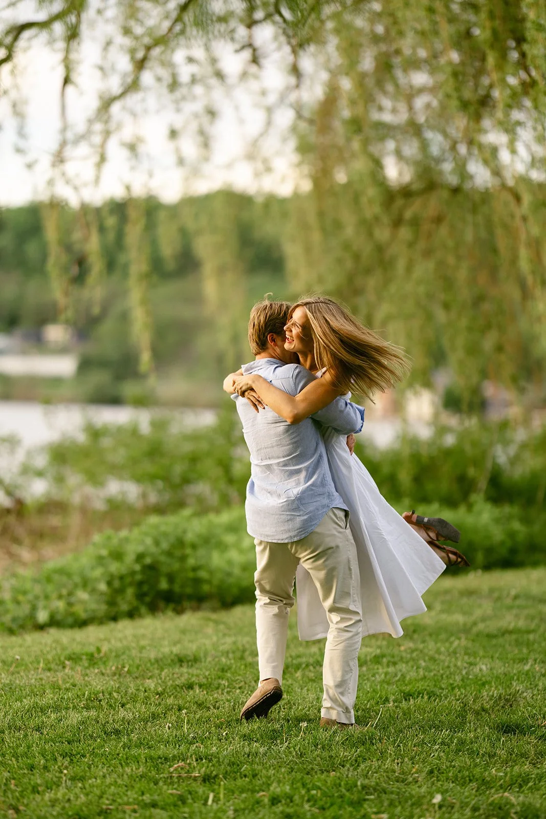 A man in light-colored clothes lifts and embraces a woman in a white dress outdoors on grass, with trees and water in the background.