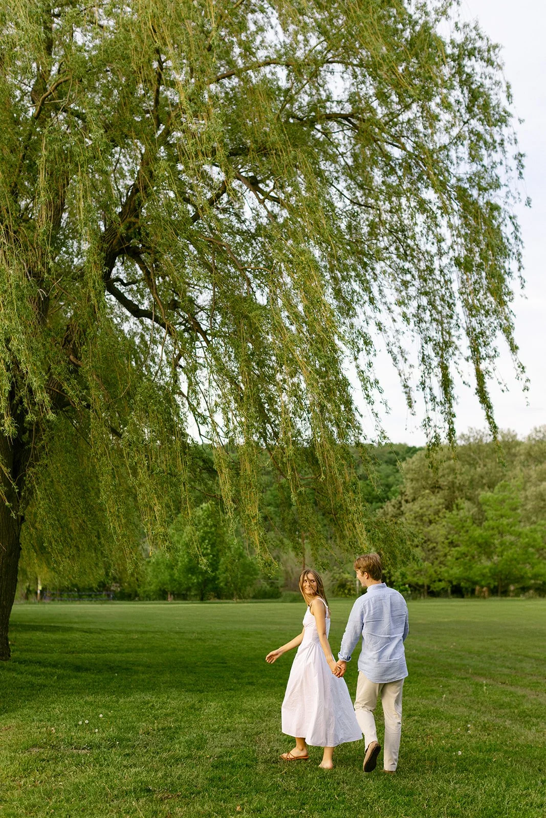A man in light-colored clothes lifts and embraces a woman in a white dress outdoors on grass, with trees and water in the background for outdoor engagement photos