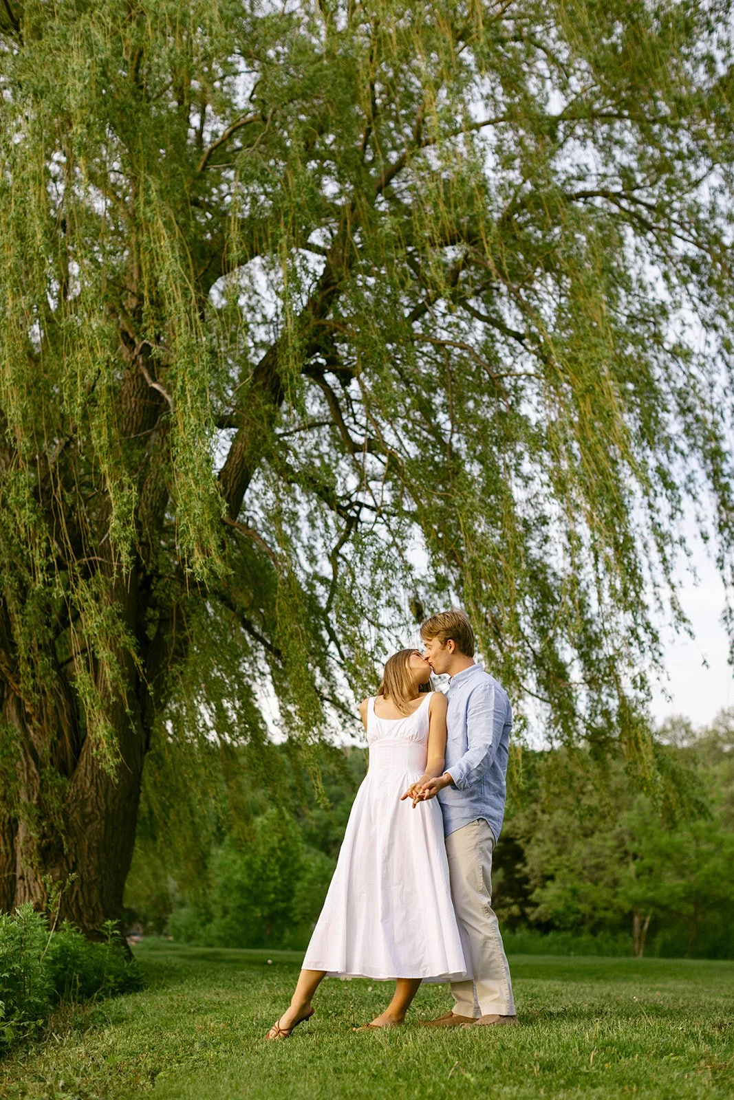 A man in light-colored clothes lifts and embraces a woman in a white dress outdoors on grass, with trees and water in the background for outdoor engagement photos