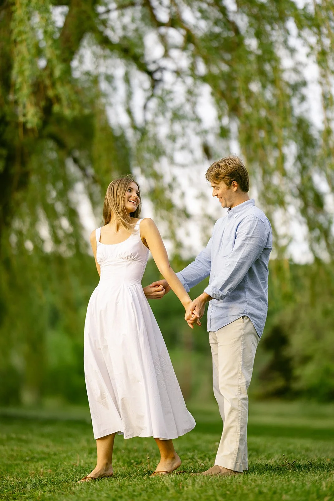 A man in light-colored clothes lifts and embraces a woman in a white dress outdoors on grass, with trees and water in the background for outdoor engagement photos
