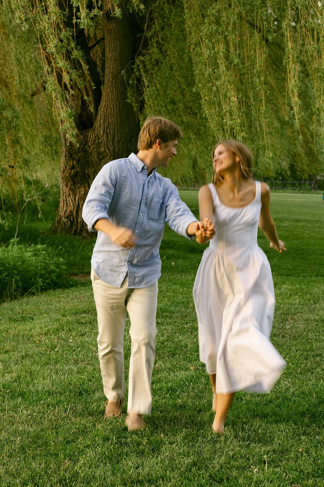 A man in light-colored clothes lifts and embraces a woman in a white dress outdoors on grass, with trees and water in the background for outdoor engagement photos