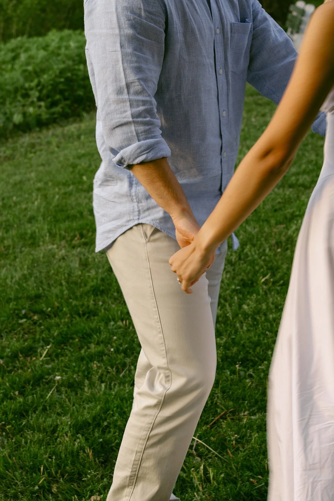 A man in light-colored clothes lifts and embraces a woman in a white dress outdoors on grass, with trees and water in the background for outdoor engagement photos