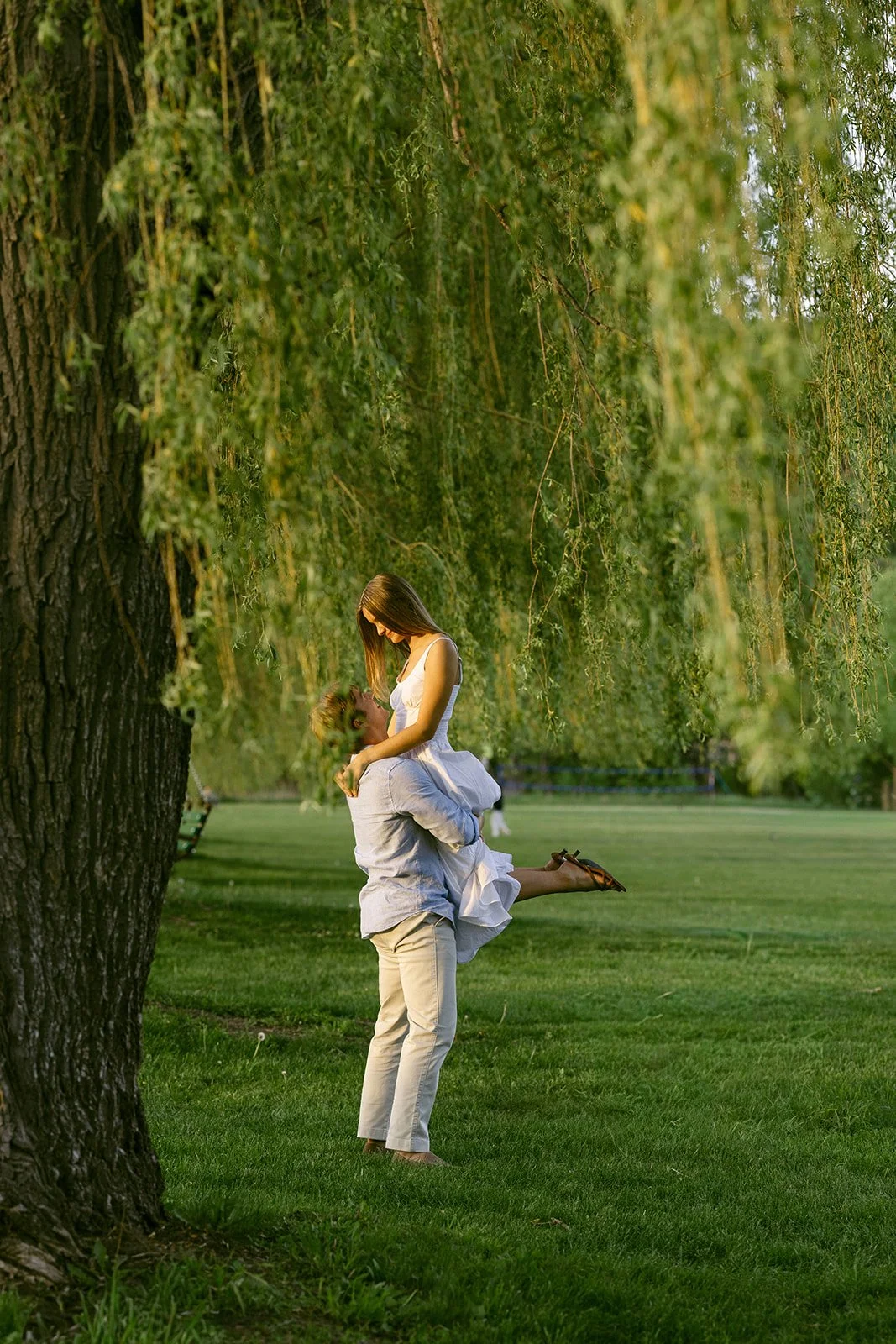A person lifts another person in a white dress under a large willow tree in a grassy park.