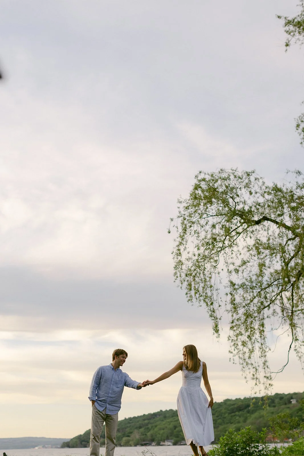 A man and woman hold hands and walk barefoot on grass near a lake, with trees and hills in the background.
