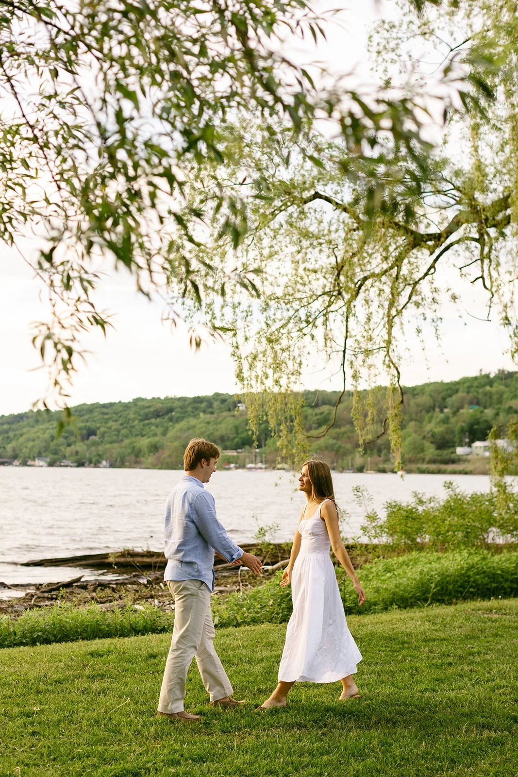 A man and woman hold hands and walk barefoot on grass near a lake, with trees and hills in the background.