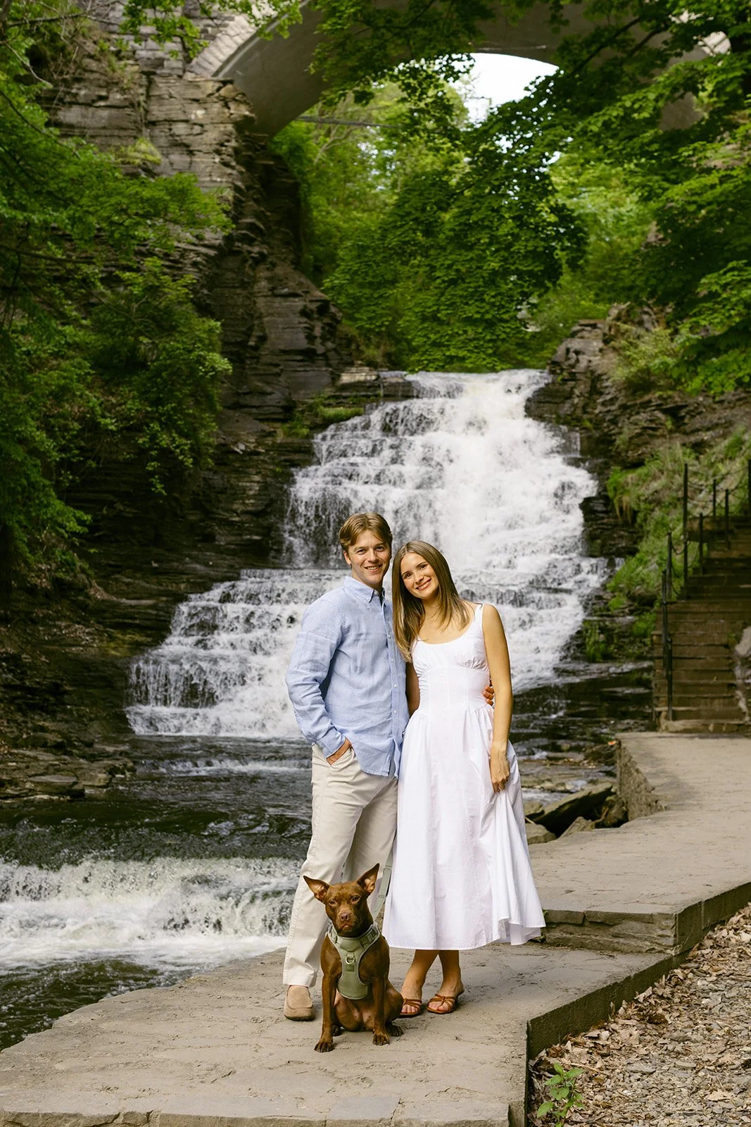 A couple stands together at the base of a multi-tiered waterfall surrounded by greenery and rocky cliffs, with water cascading down behind them for outdoor engagement photos at cascadilla gorge