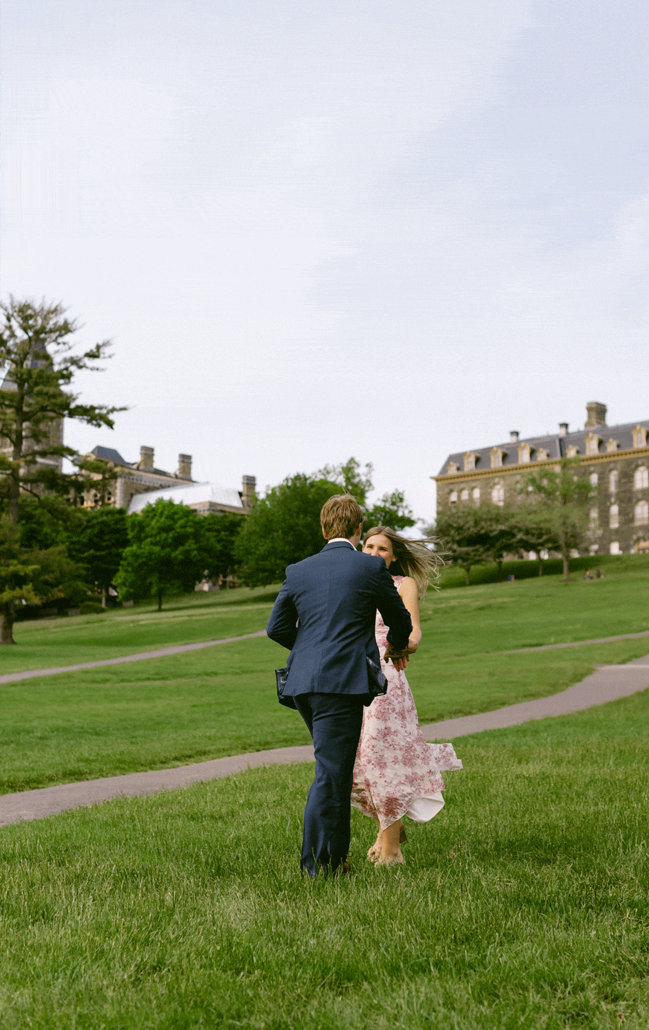 A man in a suit and a woman in a floral dress dance together on a grassy lawn, with large buildings and trees in the background.