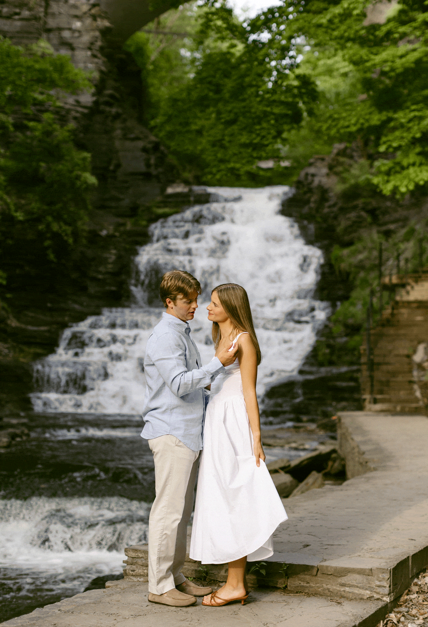 A man and woman stand close together on a stone path beside a cascading waterfall surrounded by green trees for outdoor engagement photos