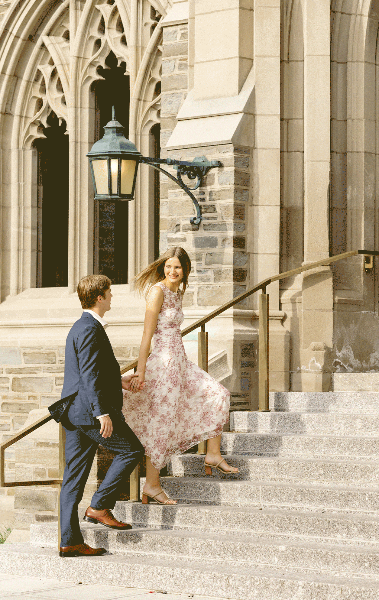 A man in a suit and a woman in a floral dress walk up stone steps outside a historic stone building with arched windows for outdoor engagement photos