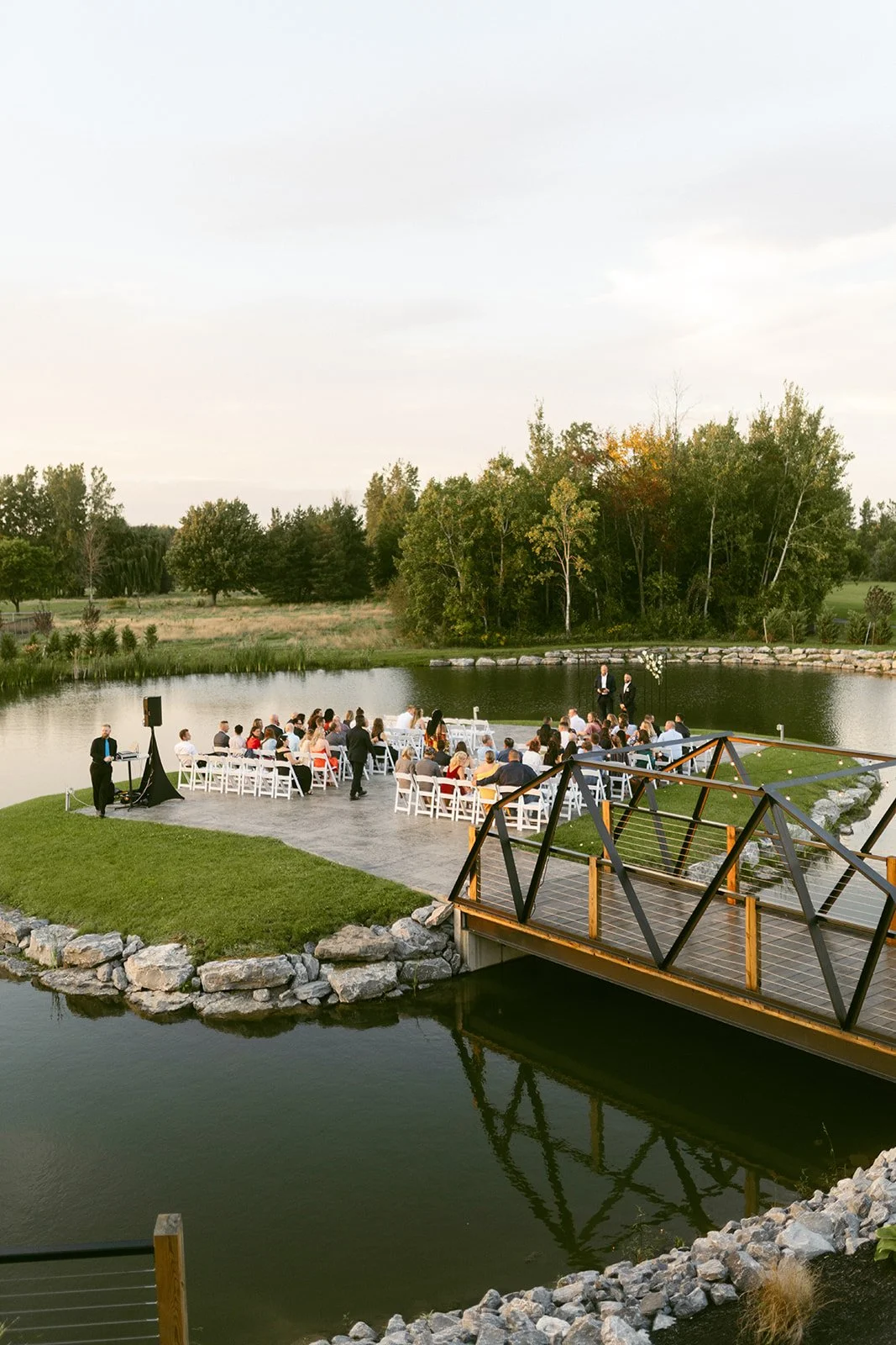 A group of people sit on white chairs by a pond for an outdoor event, with a wooden bridge in the foreground and trees in the background at the sterling at arrowhead golf club