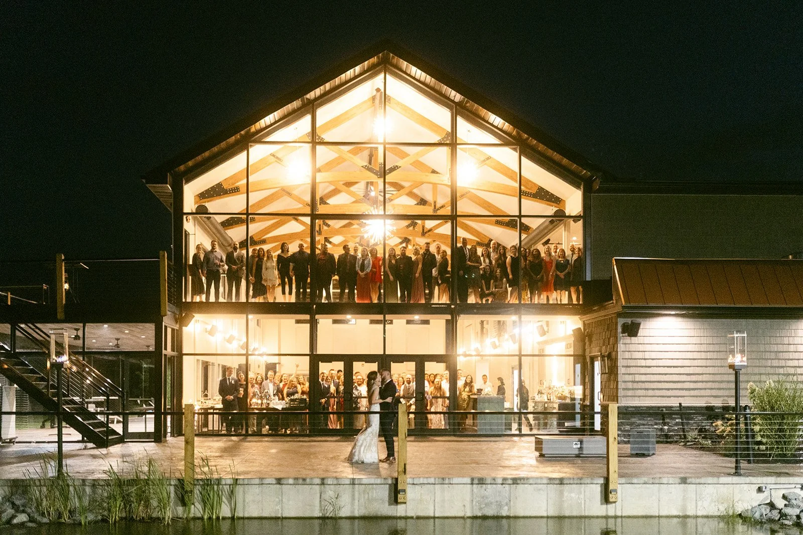 A bride and groom stand outside a modern, two-story building at night while guests gather and celebrate inside the well-lit upper floor.