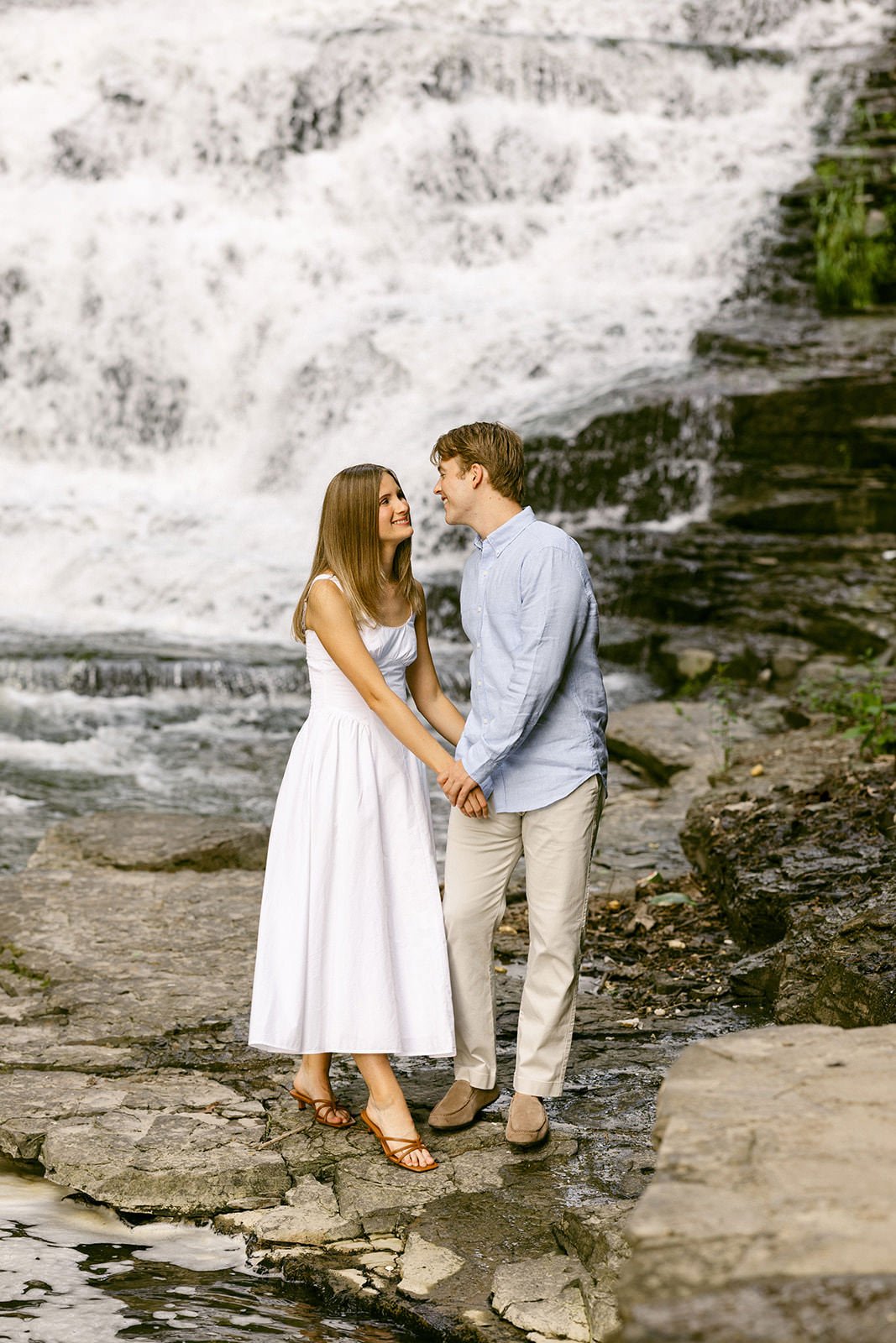 A couple stands together at the base of a multi-tiered waterfall surrounded by greenery and rocky cliffs, with water cascading down behind them for outdoor engagement photos at cascadilla gorge