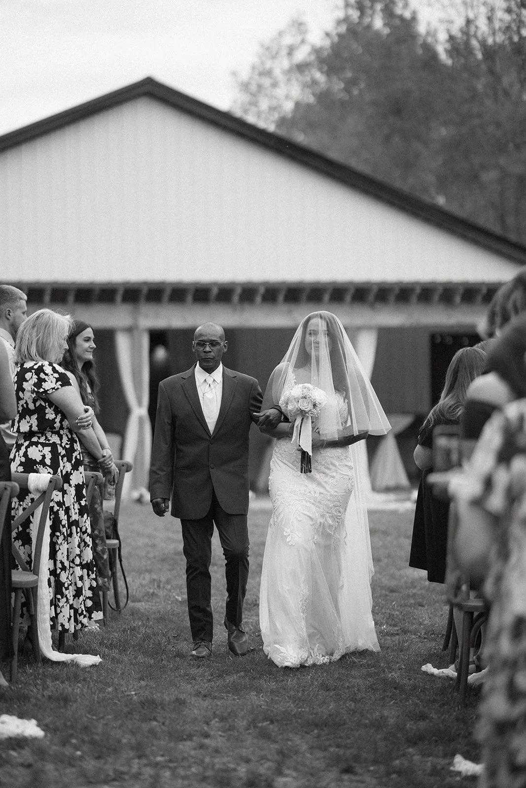 A bride in a wedding dress walks down an outdoor aisle with a man, surrounded by seated guests, in front of a barn.