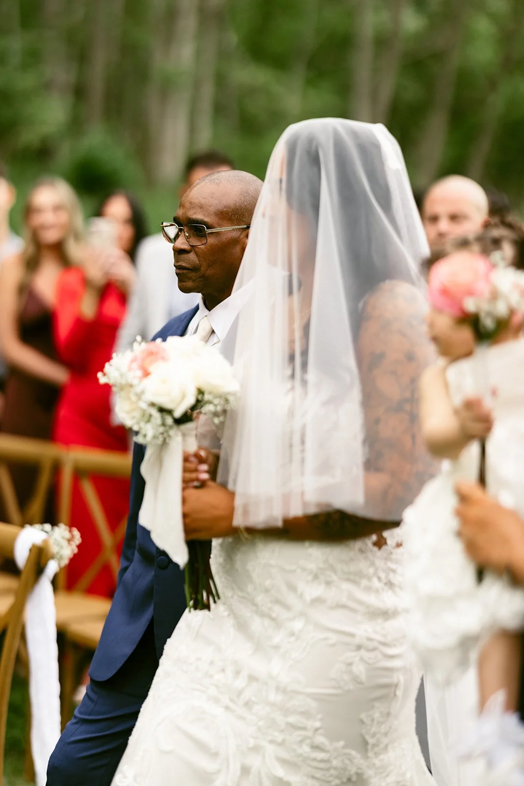 A bride in a wedding dress walks down an outdoor aisle with a man, surrounded by seated guests, in front of a barn.
